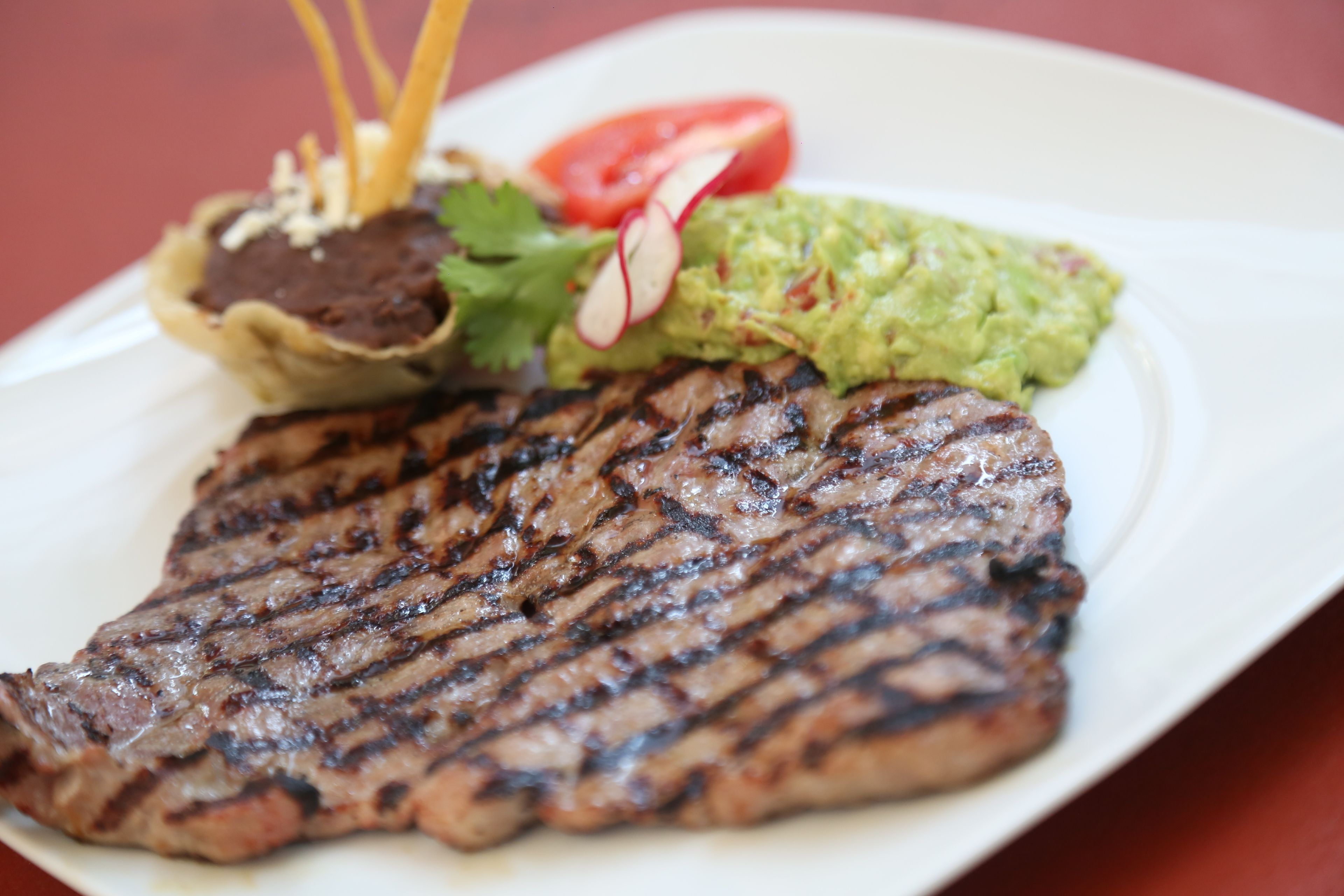Un plato con bistec a la parrilla, guacamole, una rodaja de tomate y un recipiente pequeño con salsa mole.