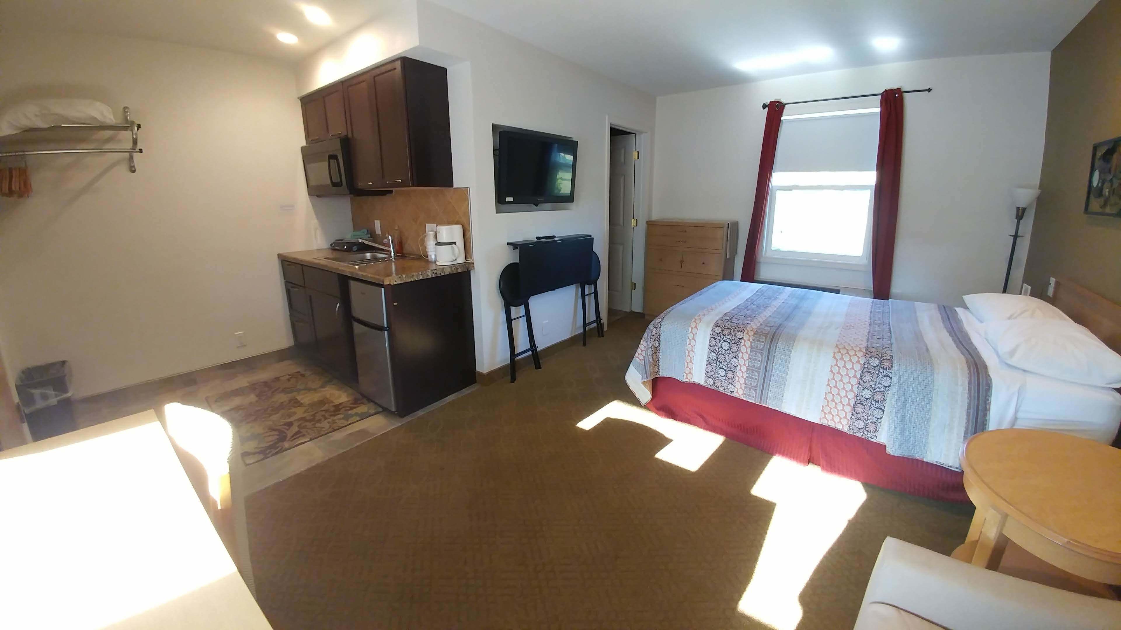 Habitación de hotel compacta con kitchenette, una cama con cubrecama estampado, un escritorio con silla y un televisor montado en la pared.