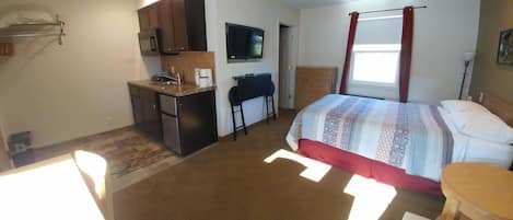 Habitación de hotel compacta con kitchenette, una cama con cubrecama estampado, un escritorio con silla y un televisor montado en la pared.