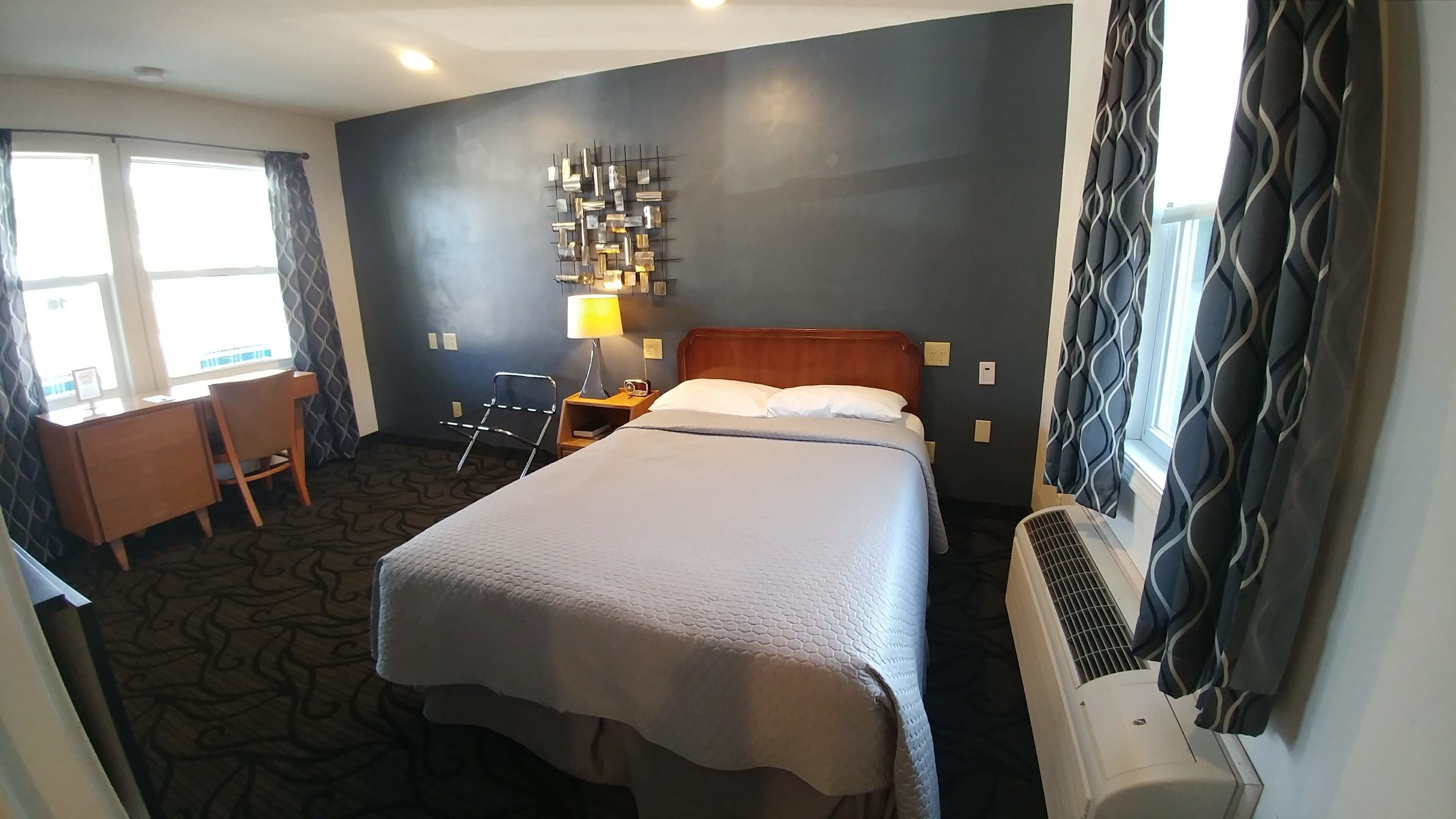 A hotel room with a bed, desk, chair, lamp, and air conditioning unit.