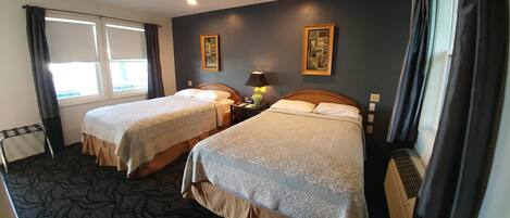 A hotel room with two beds, a window with curtains, a small table with a lamp, and a wall-mounted picture.