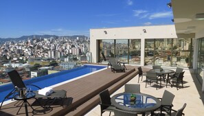 Área de piscina no terraço com espreguiçadeiras e mesas, com vista para a cidade.