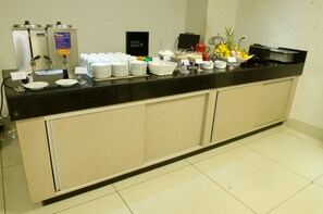 Balcão de buffet com diversas opções de comida e bebida, incluindo máquinas de café, uma cesta de frutas e uma placa com a inscrição 'matte leao'.