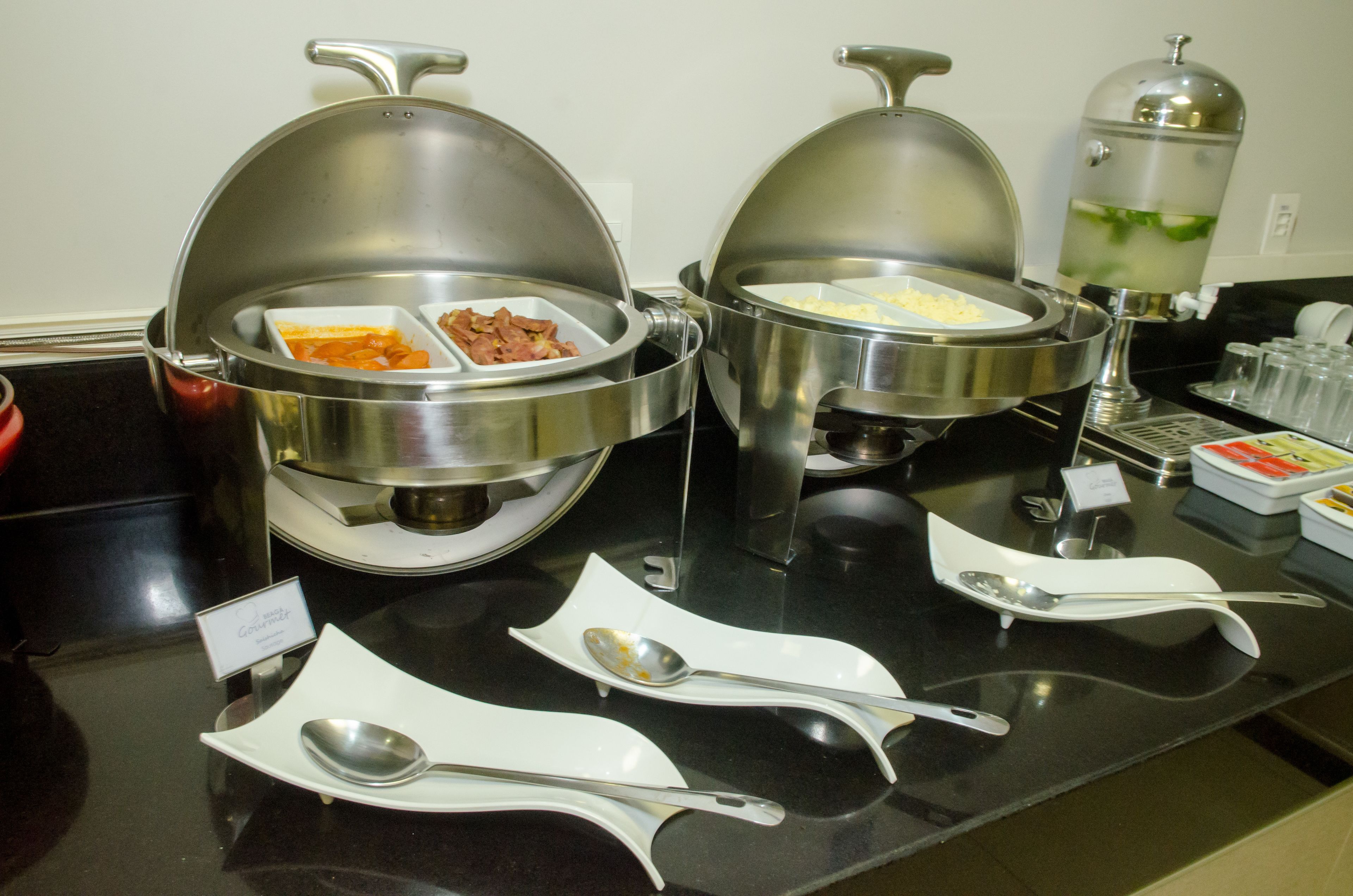 Disposición de un bufé con recipientes cubiertos para alimentos y utensilios de servicio sobre una encimera negra.