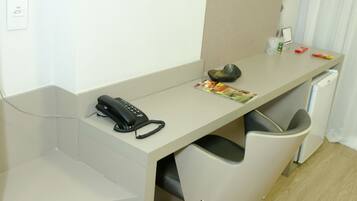 A compact hotel room with a desk, a telephone, a trash bin, and a small refrigerator.
