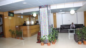 Zona de recepción con un mostrador de madera, un reloj y un cartel vertical colorido. Se observan plantas en macetas y un sofá al fondo.