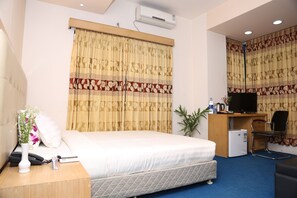 A hotel room with a bed, bedside table, a desk with a computer, a chair, and an air conditioning unit.