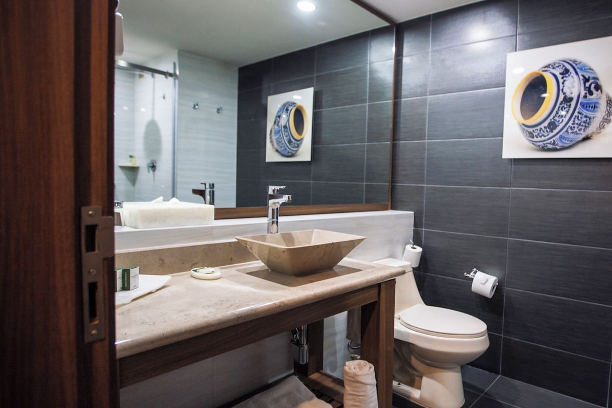A modern bathroom with a large mirror, a vessel sink, a toilet, and dark tiled walls.