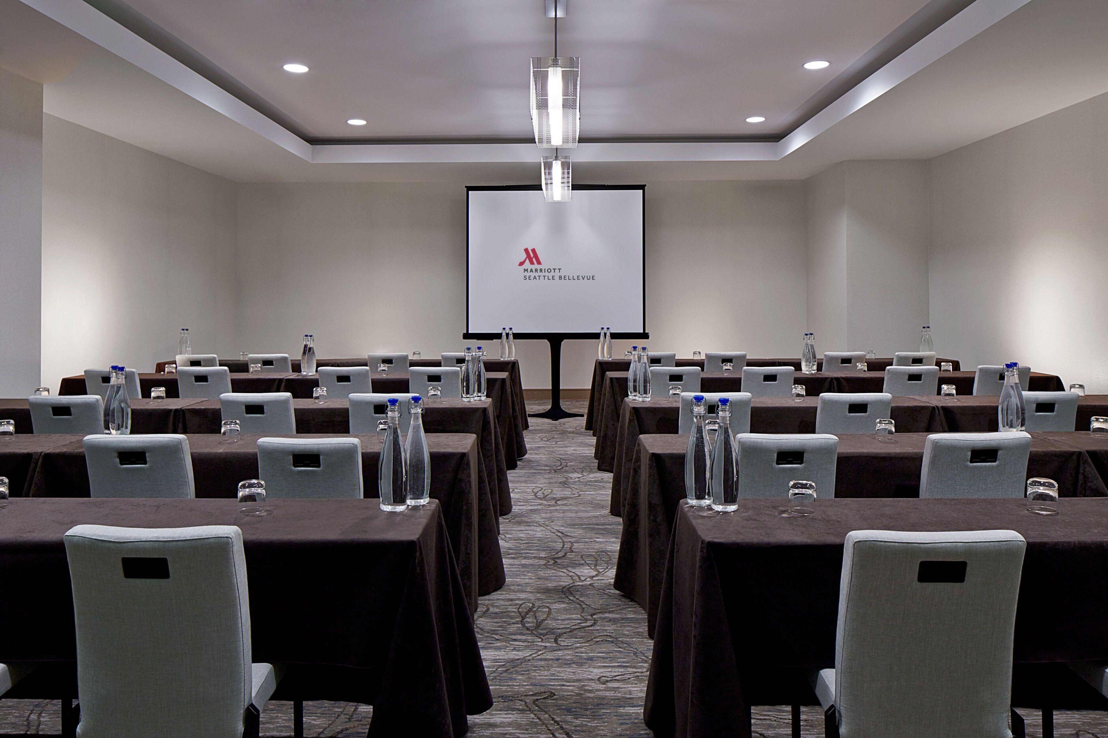 A conference room with multiple tables, each with water bottles and glasses, and a large screen displaying the Marriott Seattle Bellevue logo.