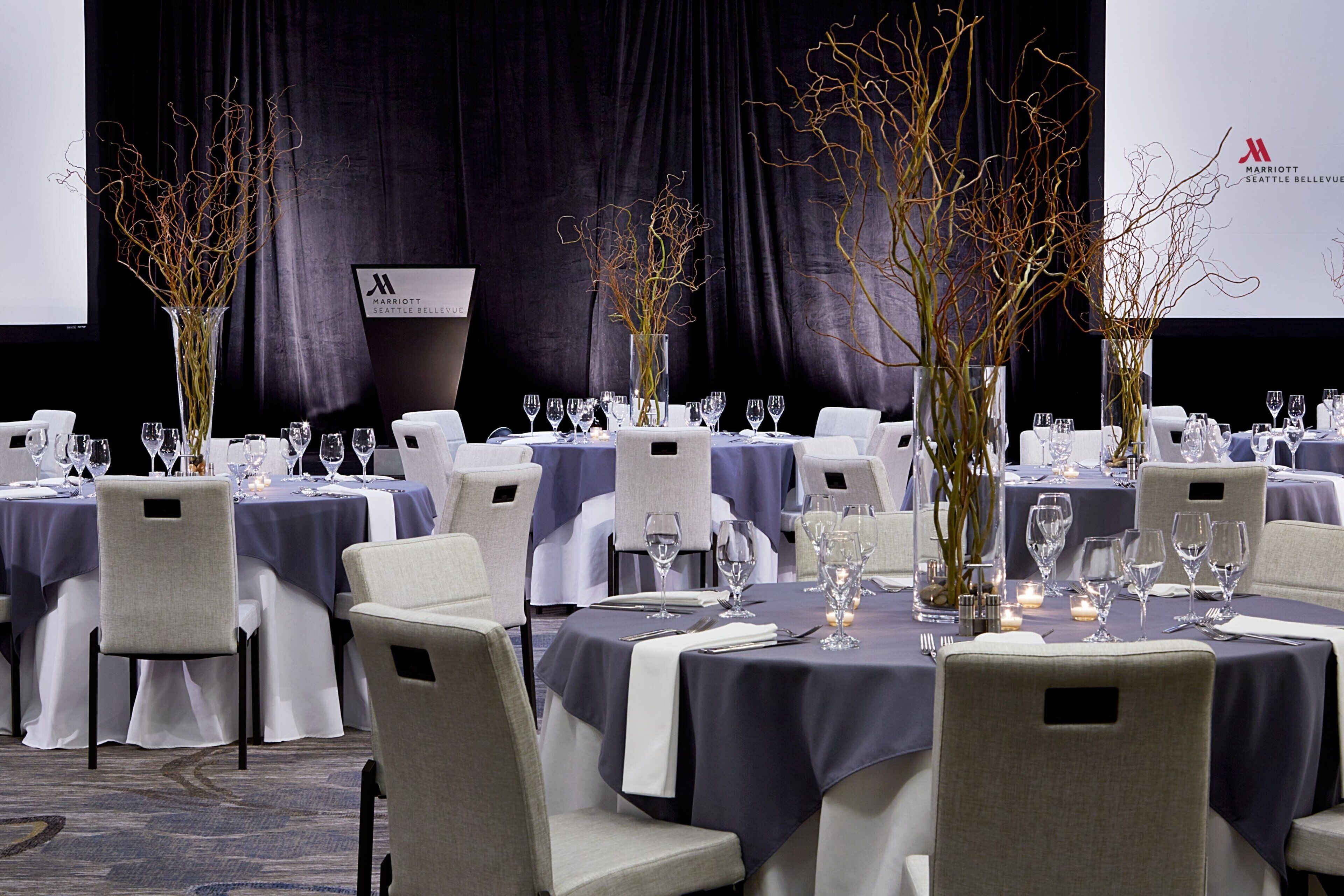 Un salón de banquetes con mesas redondas dispuestas para cenar, cada una con mantel blanco y cubiertos.
