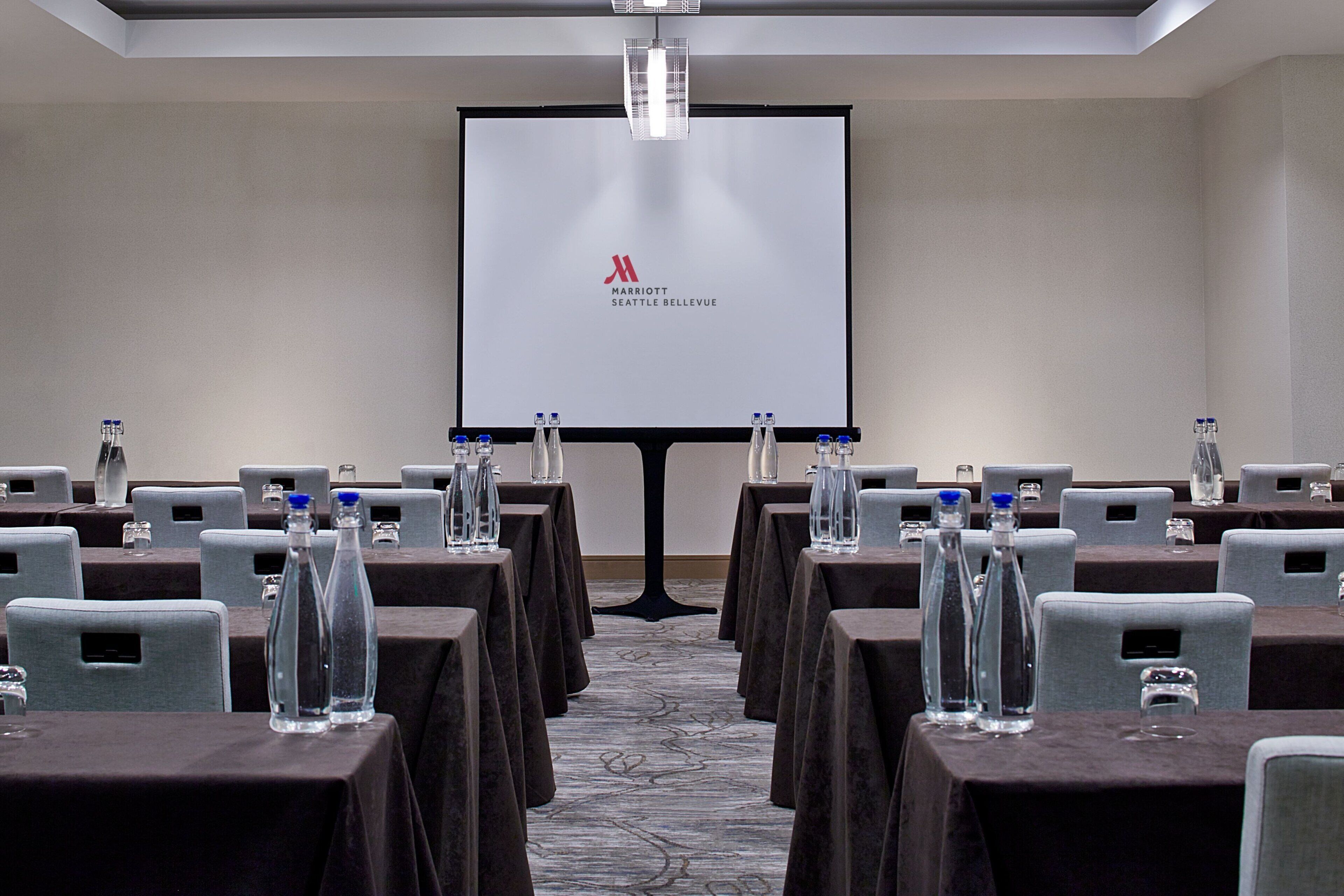 A conference room with rows of tables, each with a water bottle and a glass. A projector screen displays the Marriott Seattle Bellevue logo.