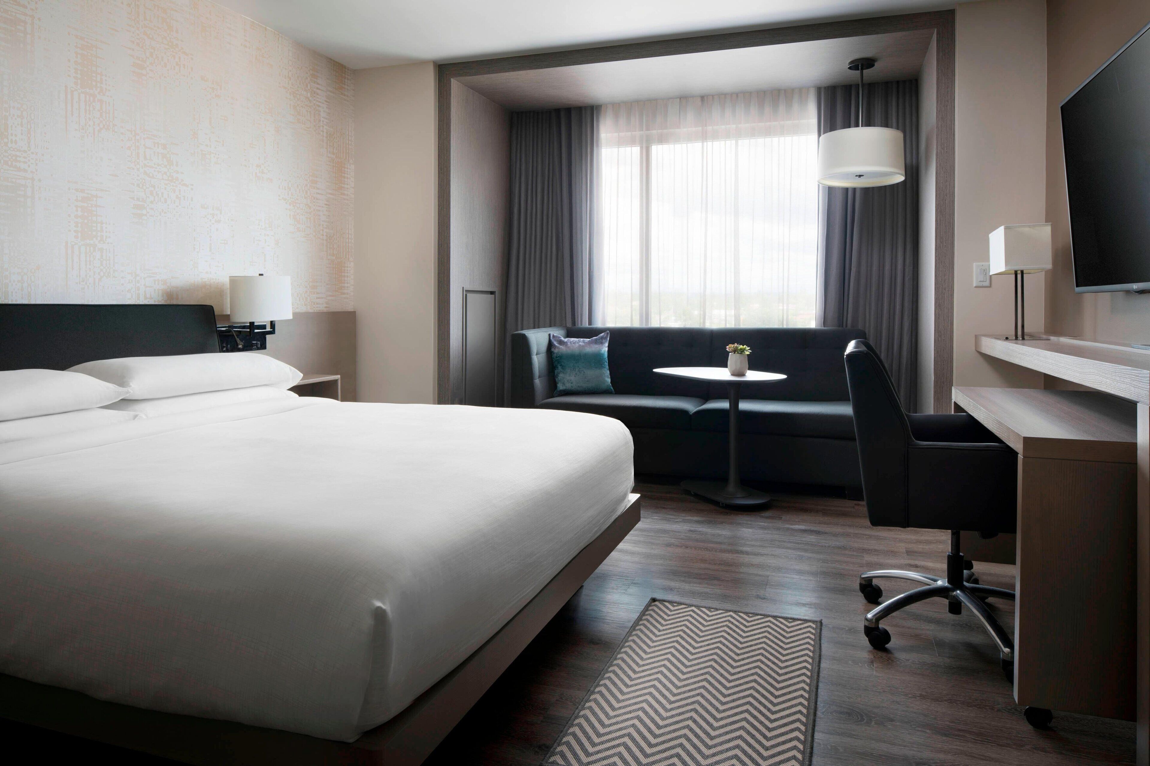 A hotel room with a large bed, a desk, a chair, a small table, a sofa, and a TV.