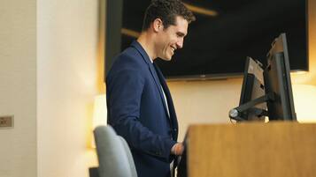 Un homme en costume bleu se tient devant un bureau, avec un écran d’ordinateur et un téléviseur fixé au mur.