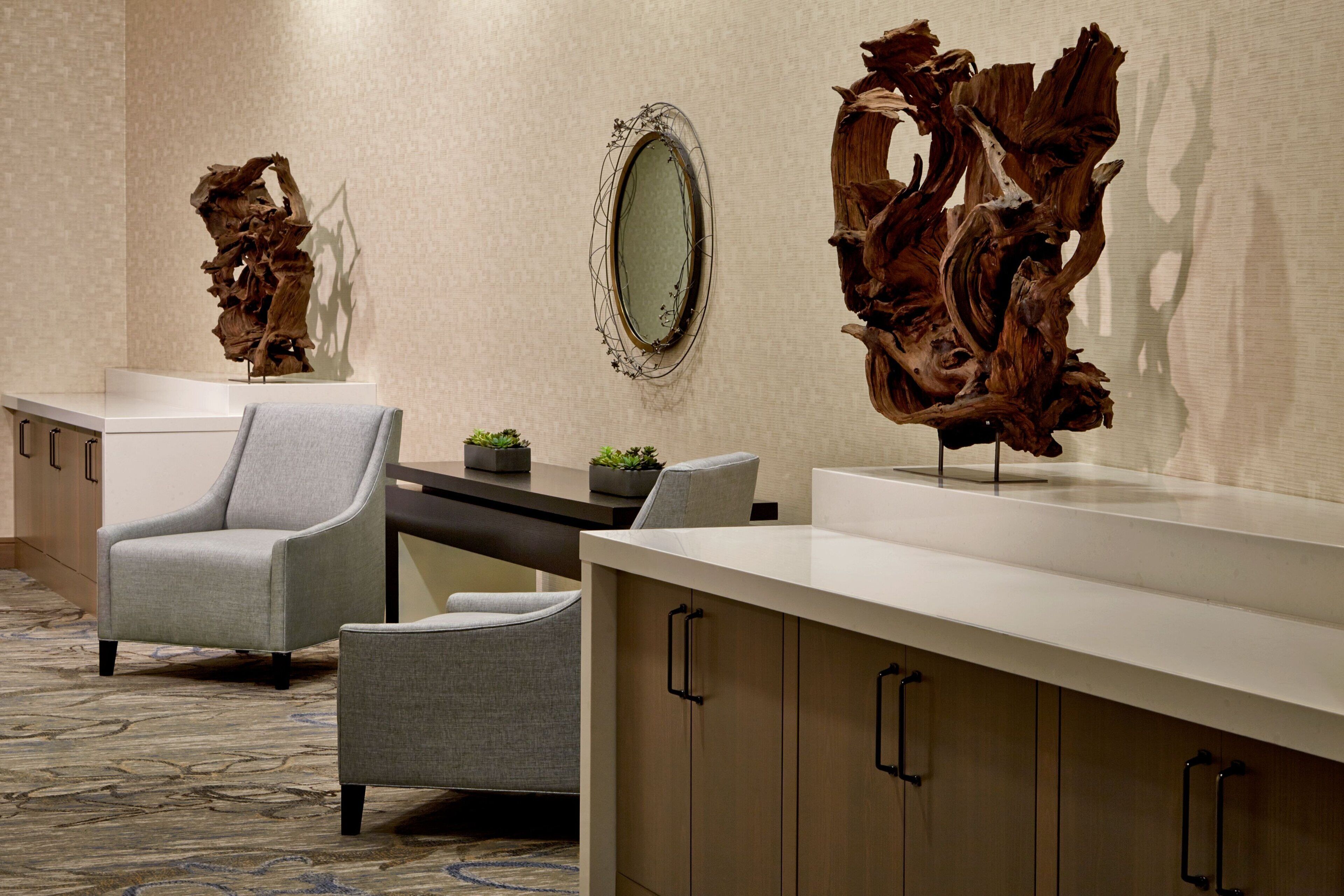 A modern hotel lobby with a reception desk, two grey armchairs, a small potted plant, and a large abstract wooden sculpture.