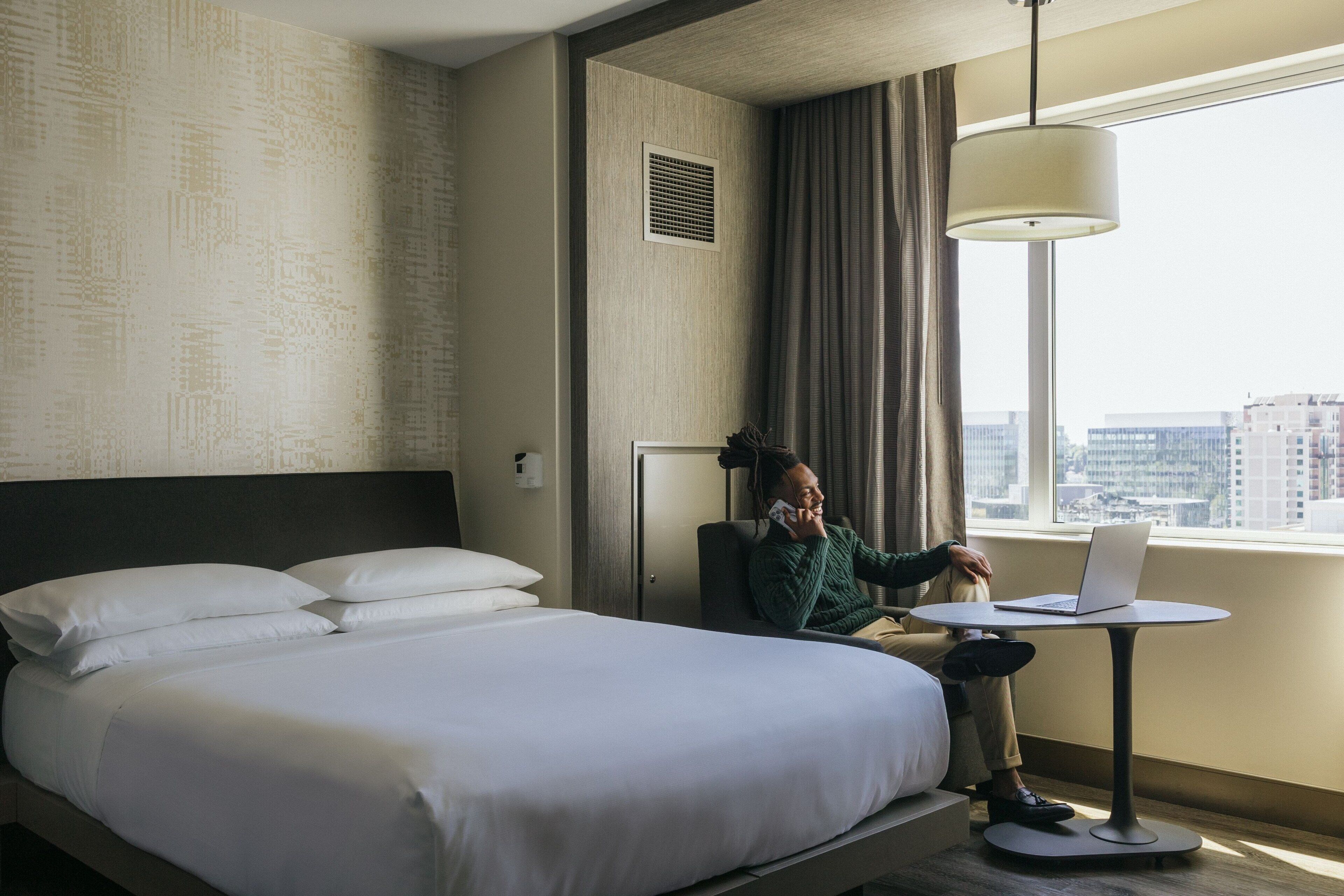 A hotel room with a bed, a desk with a laptop, a window with a city view, and a wall-mounted vent.