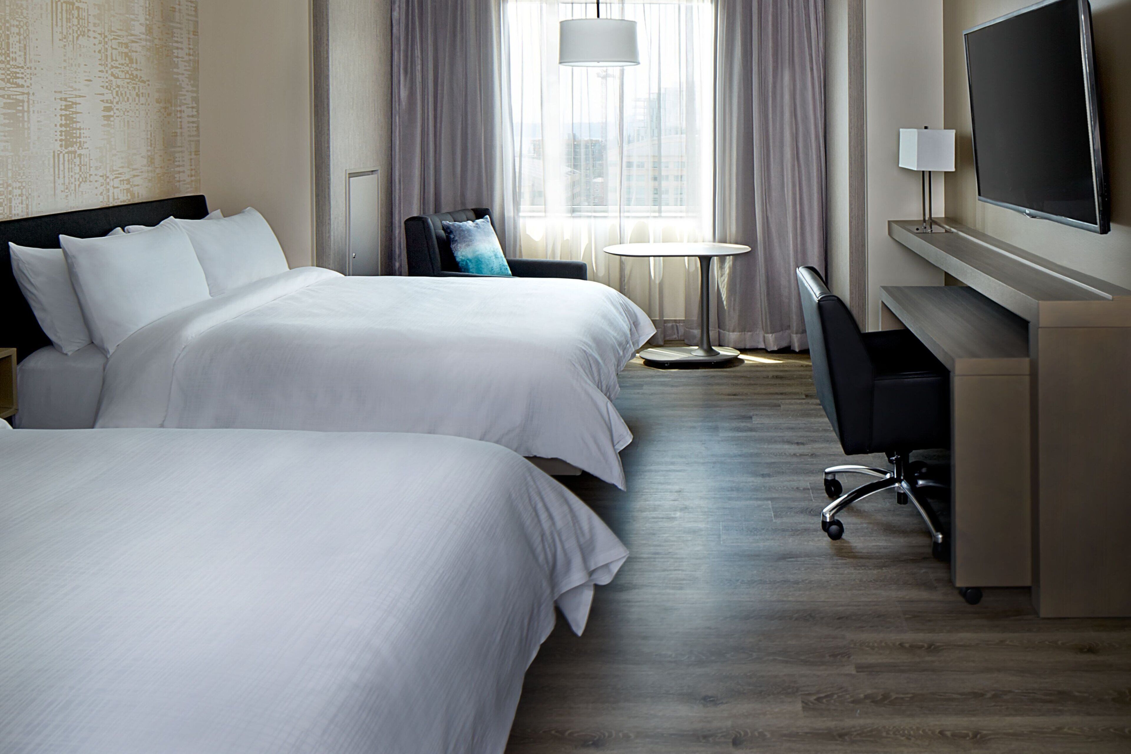 Quarto de hotel com duas camas, uma mesa, uma cadeira, uma televisão e uma janela com cortinas.
