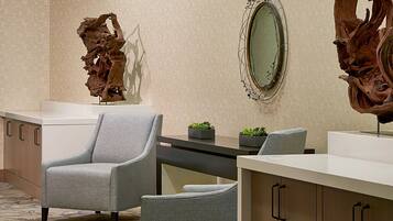 Un moderno hall de hotel con un sillón gris, una escultura de madera, un espejo y una planta pequeña en la recepción.