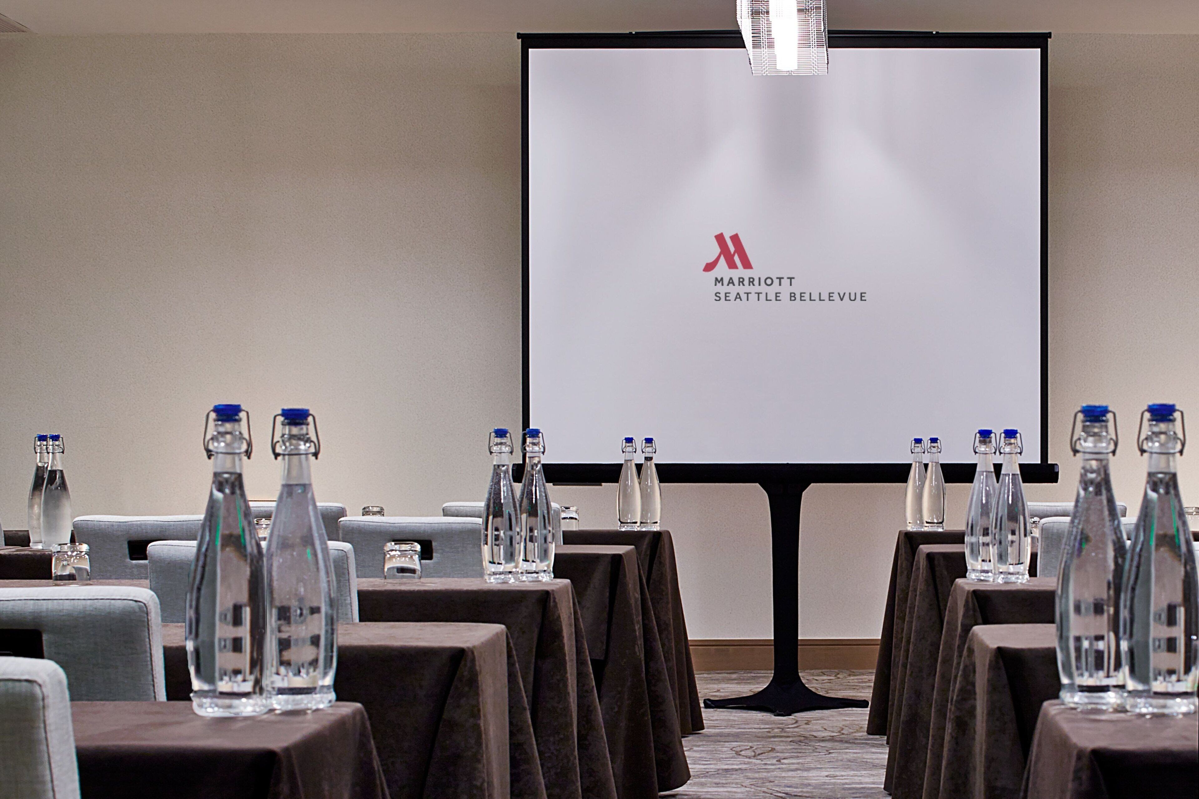 Un salón de conferencias con mesas dispuestas para una reunión, cada una con botellas de agua y vasos. Una pantalla grande muestra el logotipo de Marriott Seattle Bellevue.