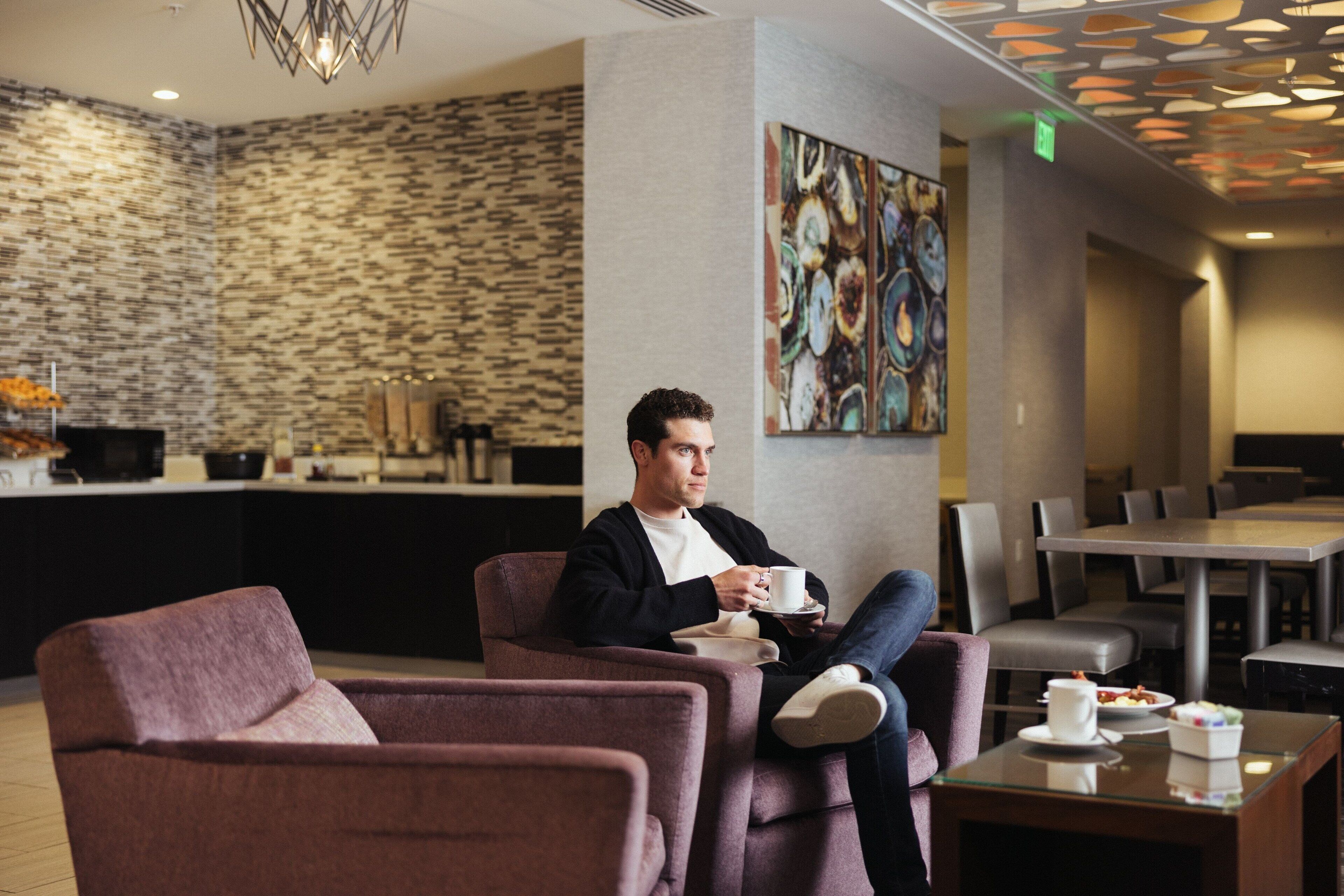 A man sitting in a lounge chair with a cup, a dining area with tables and chairs, and a patterned wall in the background.