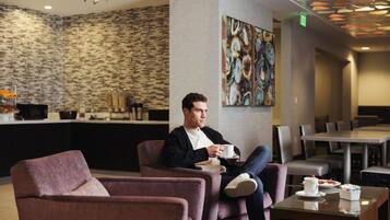 A man sitting in a lounge chair with a cup, a dining area with tables and chairs, and a patterned wall in the background.