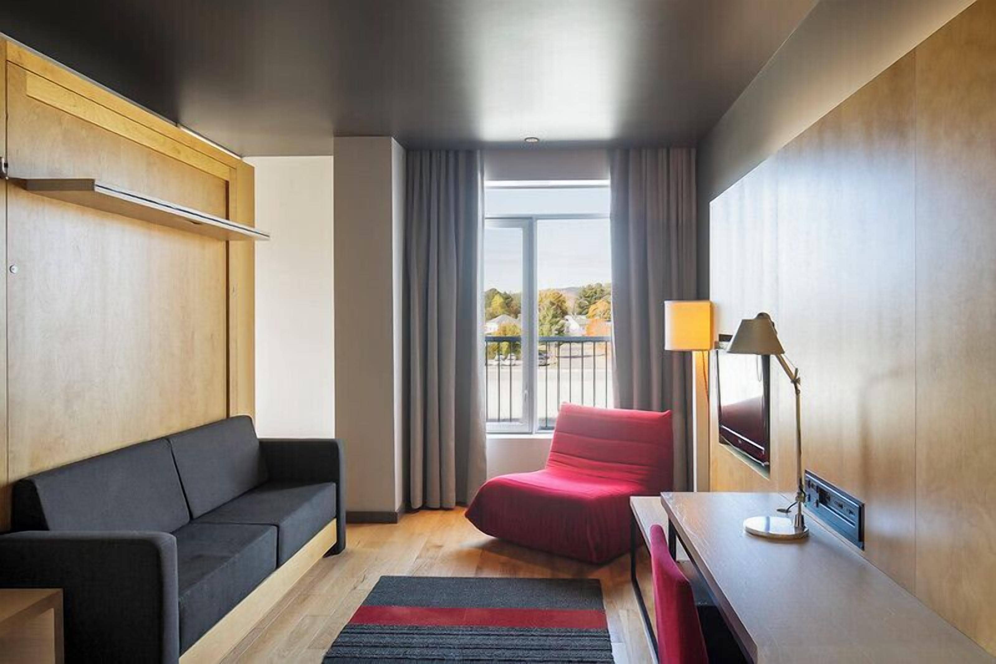 A modern hotel room with a sofa, a red chair, a desk with a lamp, and a large window with curtains.