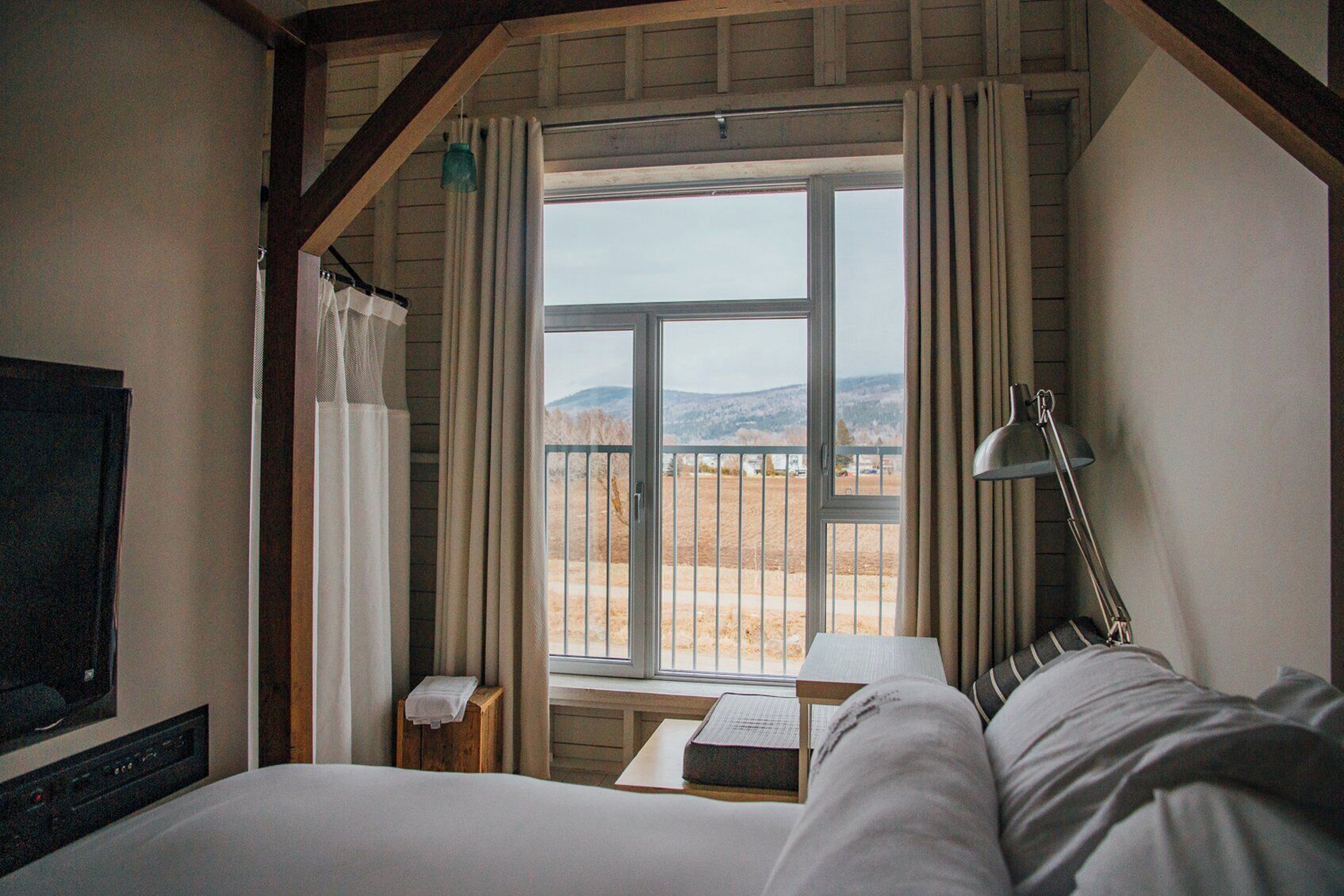 A bedroom with a large window, a bed, a bedside table, a lamp, and a TV mounted on the wall.
