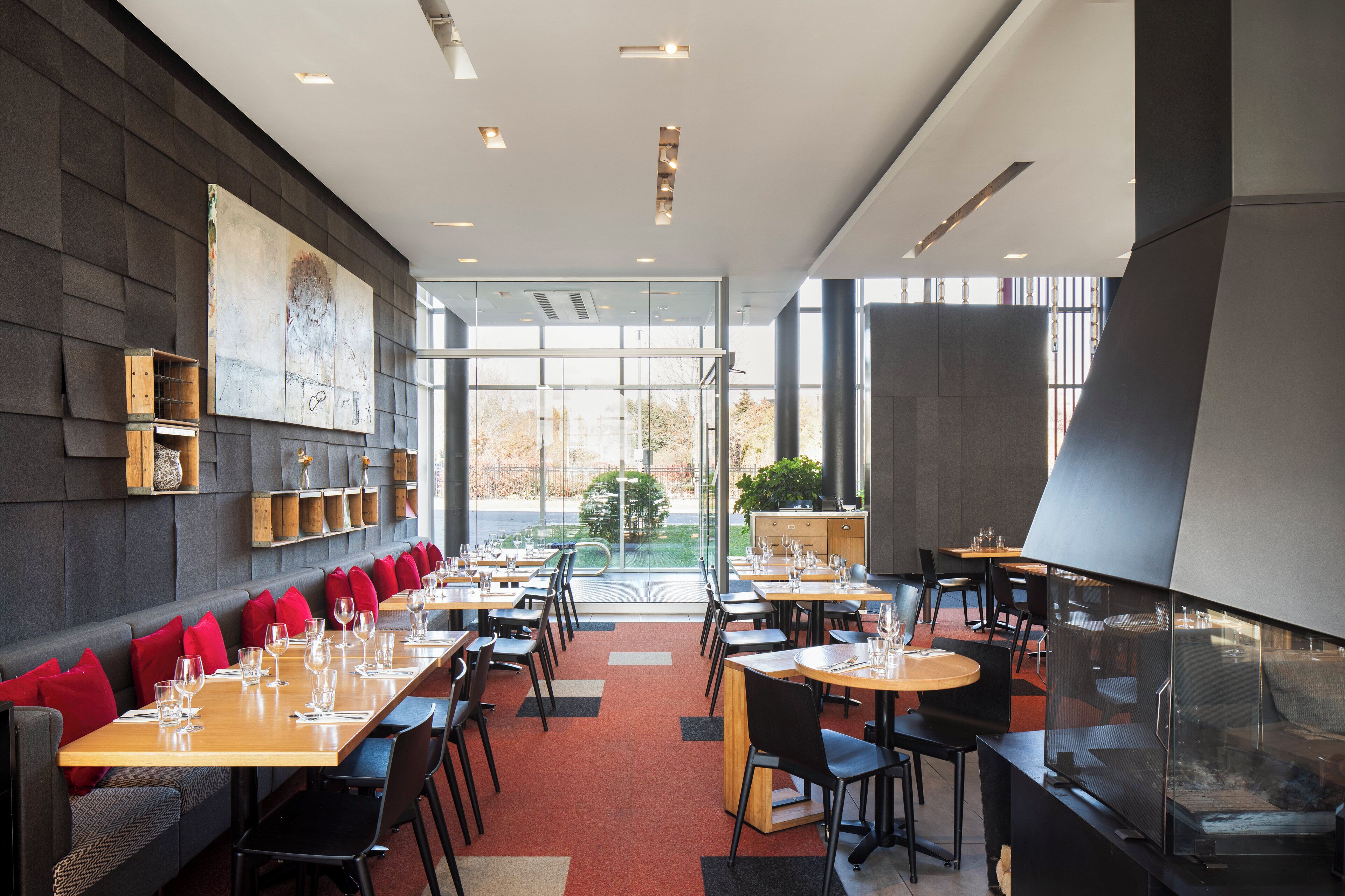A modern restaurant with long tables, red cushioned seating, and a large window offering a view of the outdoors.