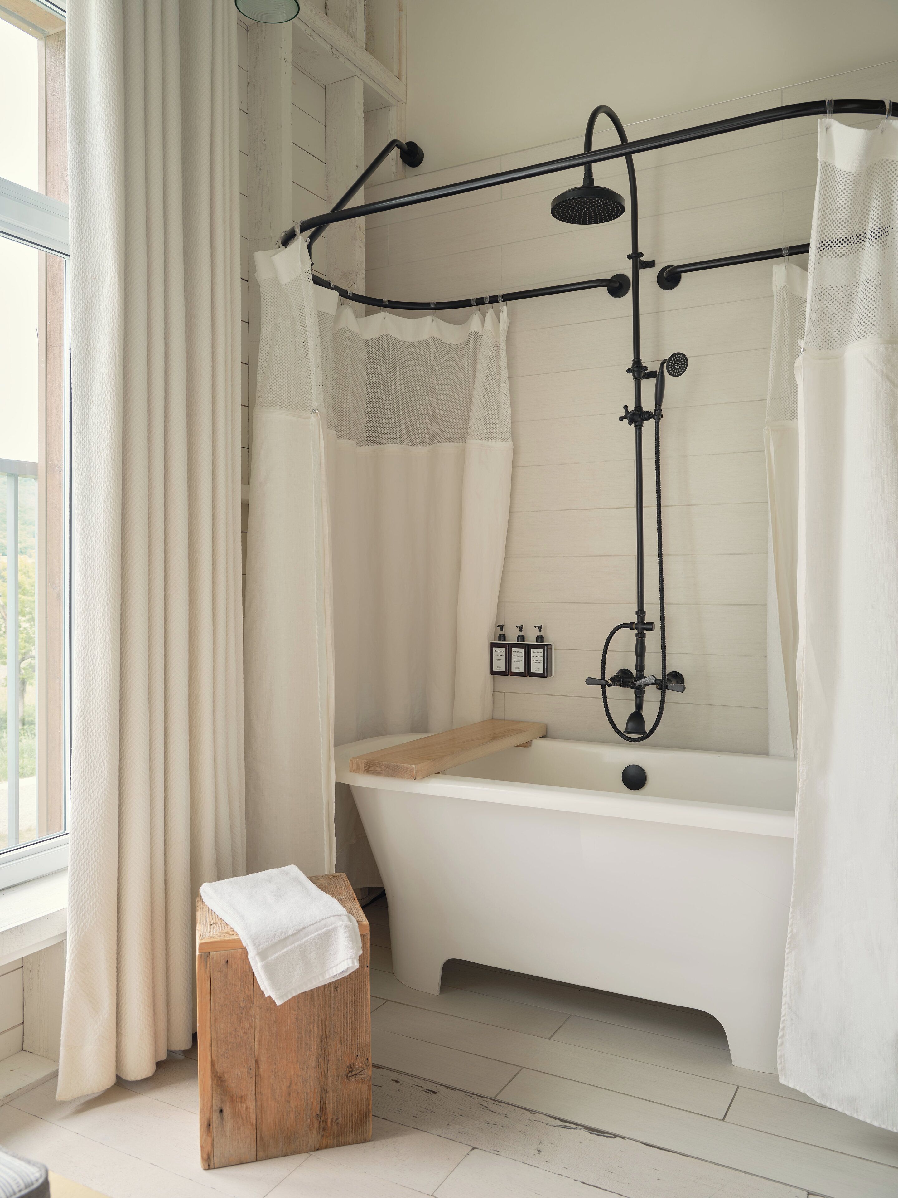A modern bathroom with a freestanding bathtub, a wooden stool, and a shower with a black metal frame.