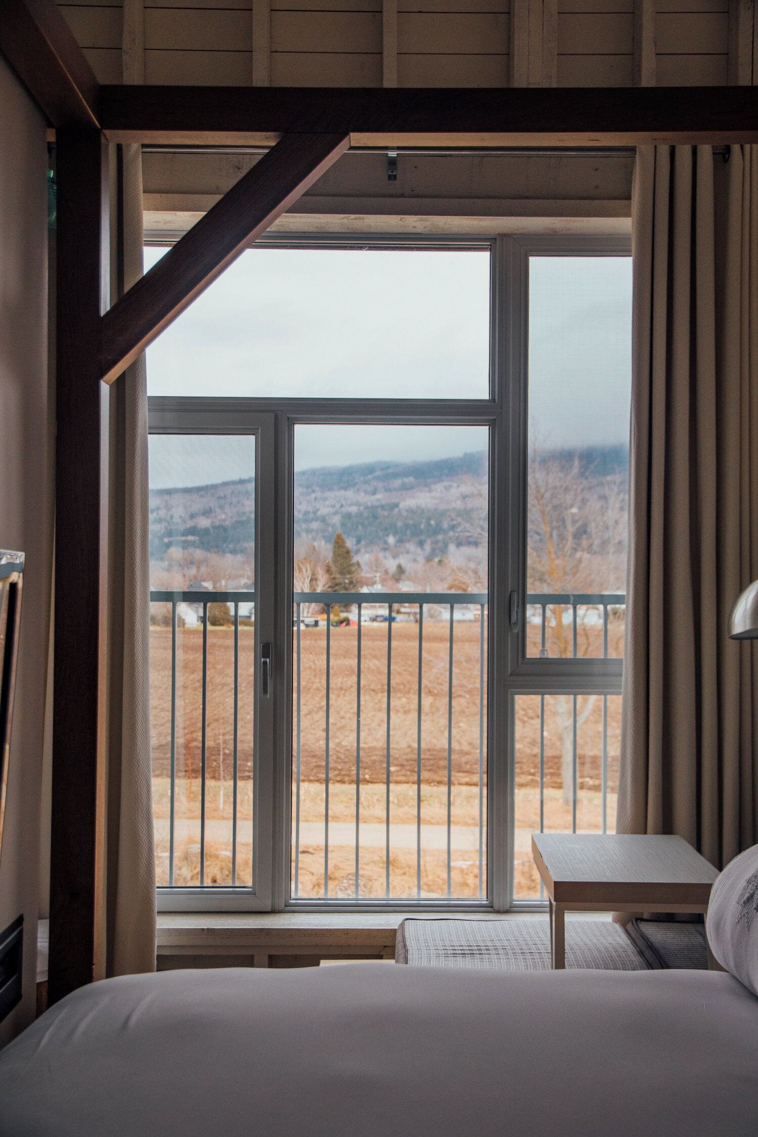Une chambre avec une grande fenêtre, un lit, une table de chevet et une vue sur un paysage montagneux.