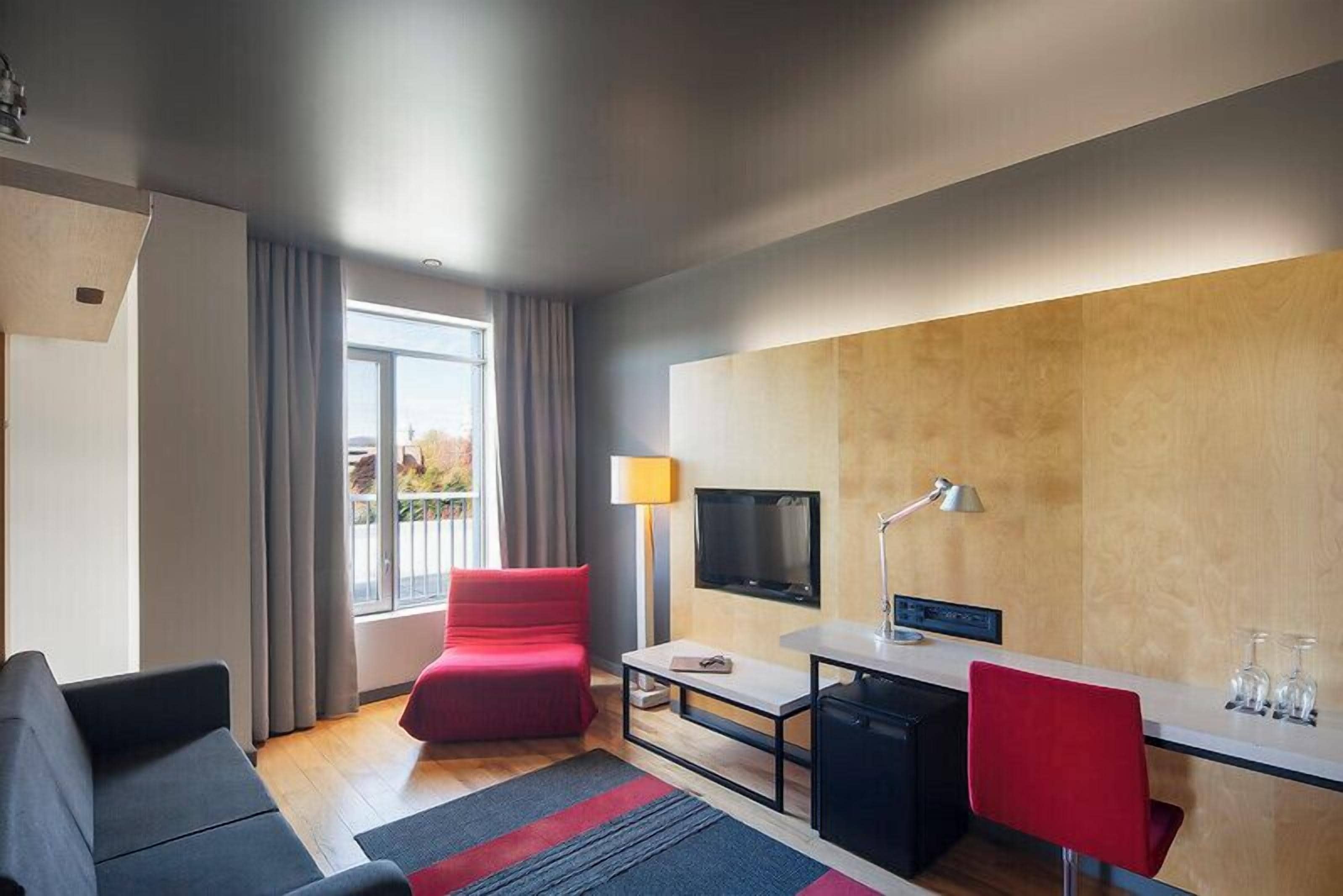 Une chambre d’hôtel moderne avec un canapé, un fauteuil rouge, une télévision, un bureau et une vue par la fenêtre.