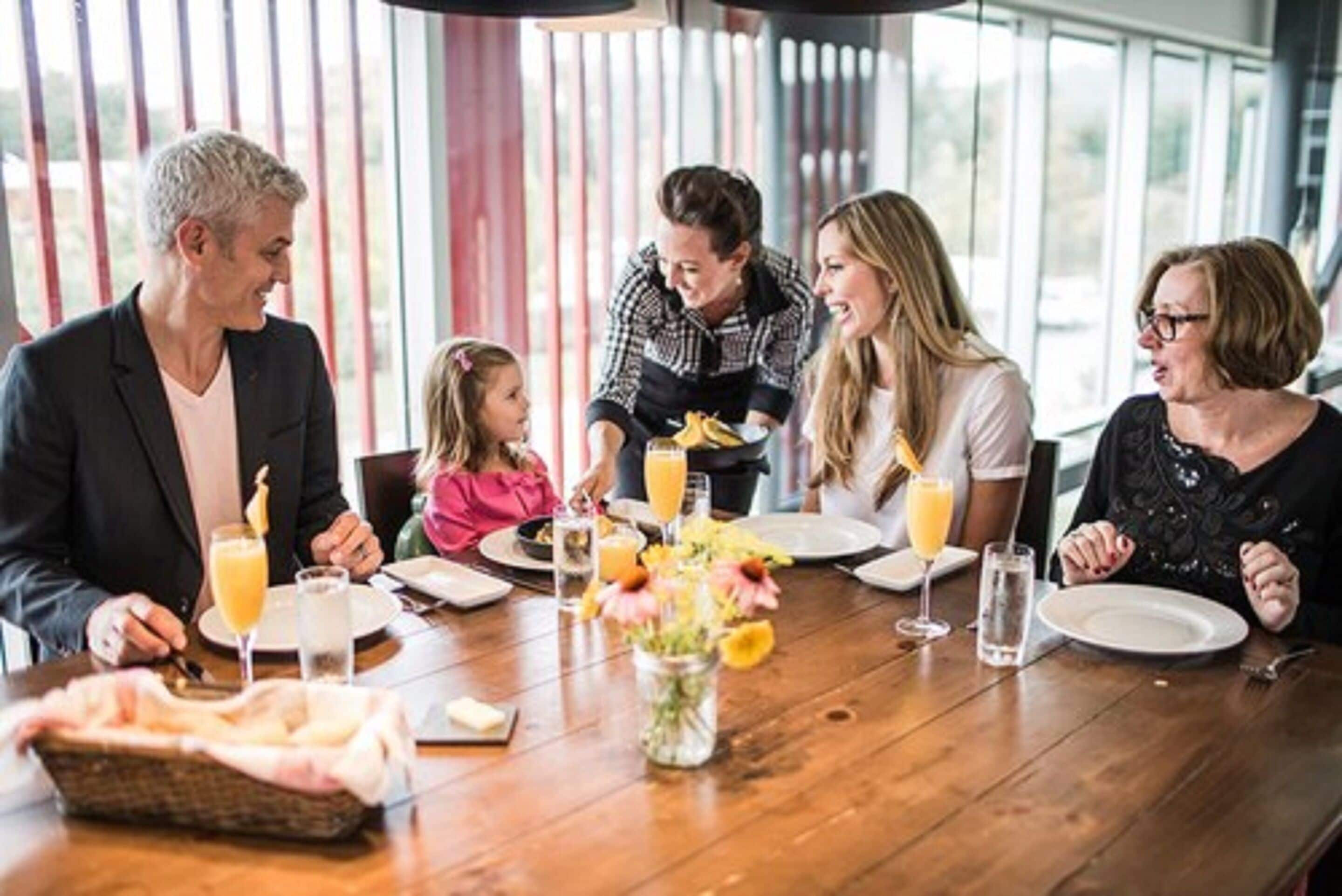 Una familia cenando en una mesa de madera con una cesta de pan, un jarrón de flores y bebidas.