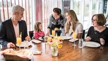 Une famille est assise autour d’une table en bois, devant un panier de pain, un vase de fleurs et des boissons.