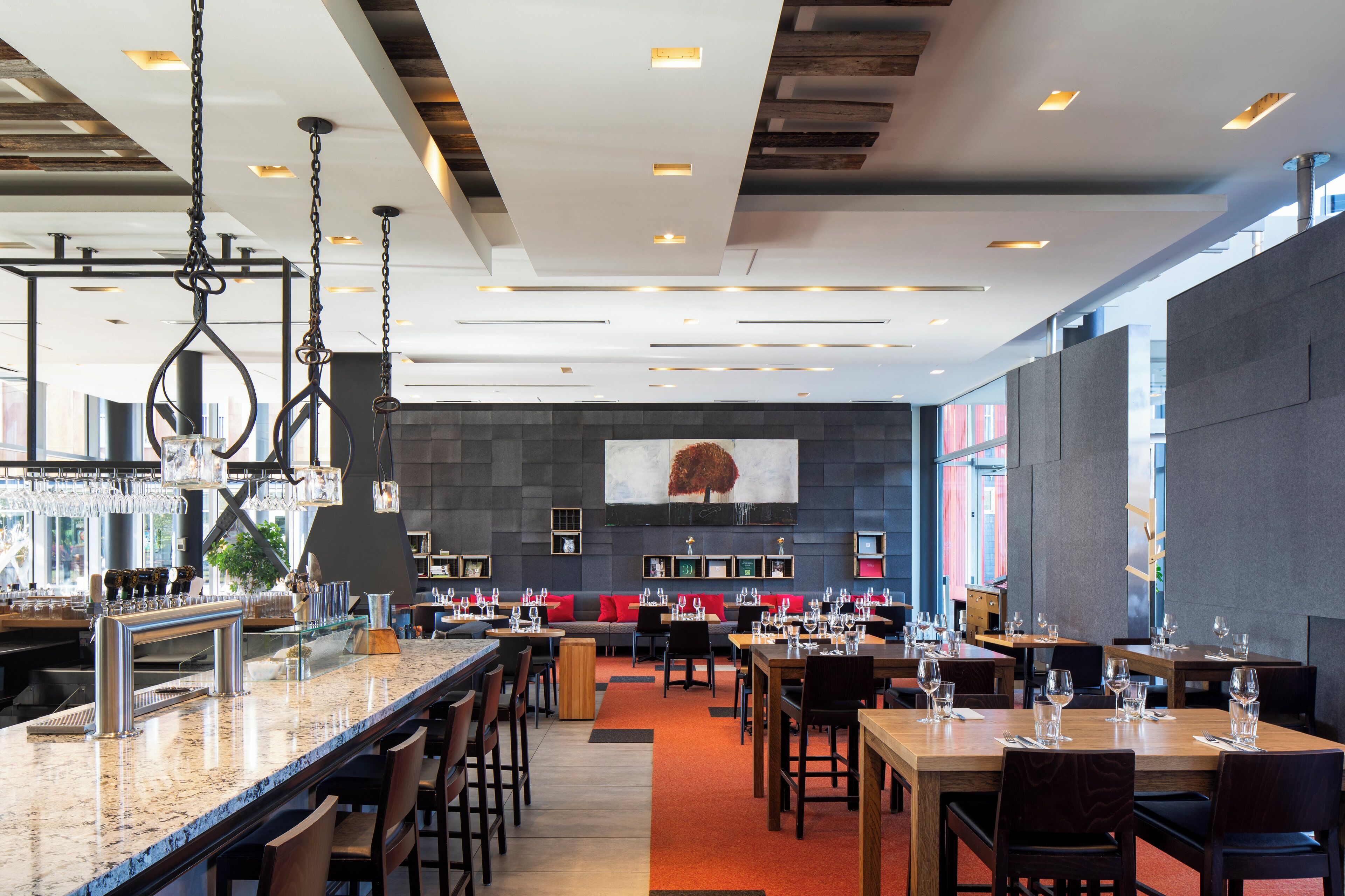 Interior de un restaurante moderno con barra, diferentes disposiciones de asientos y una gran obra de arte en la pared.