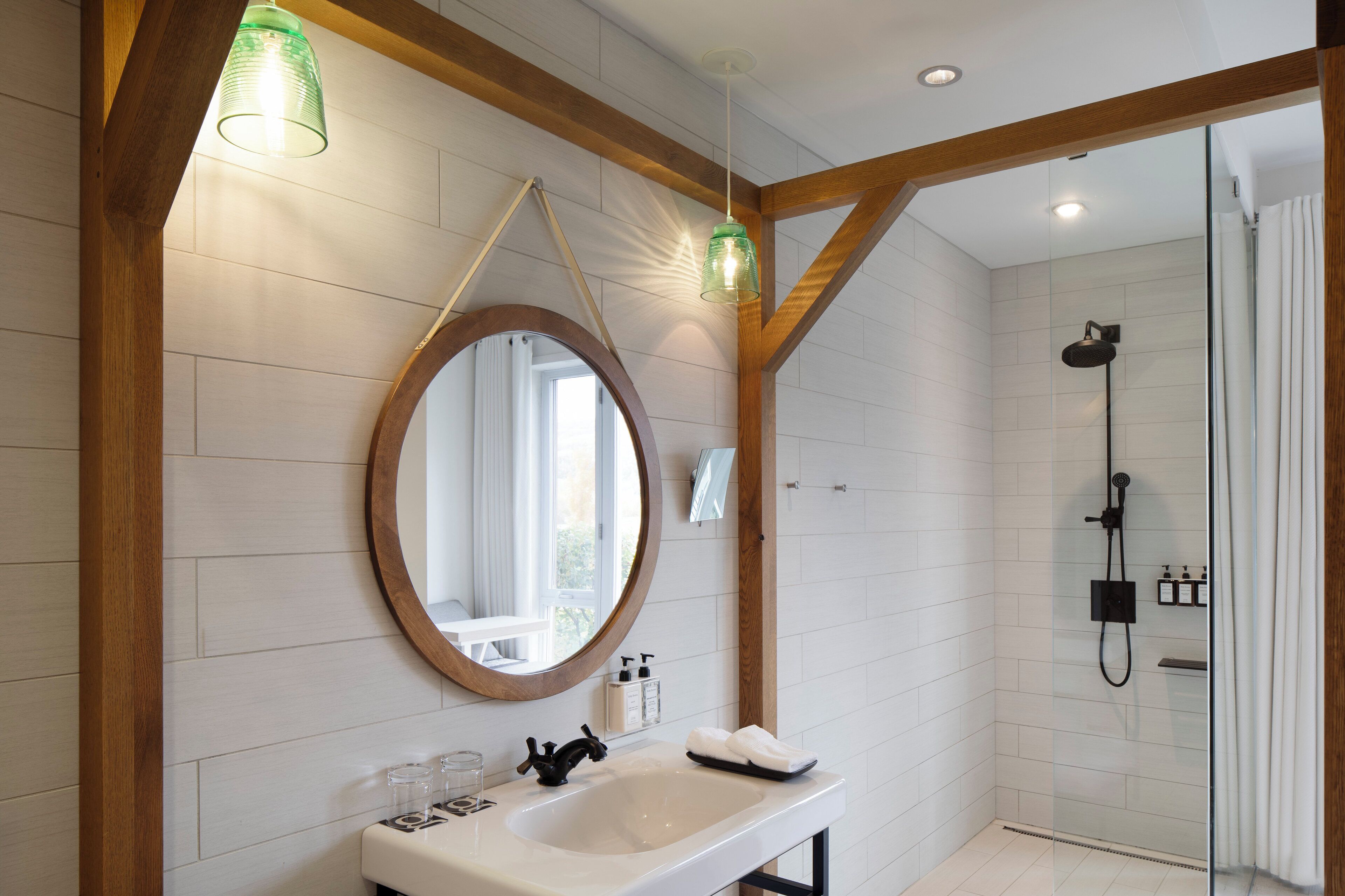 A modern bathroom with a wooden beam ceiling, a round mirror, a white sink, and a glass-enclosed shower area.