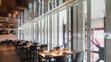 A modern restaurant with long tables, chairs, and a high ceiling with exposed ductwork.