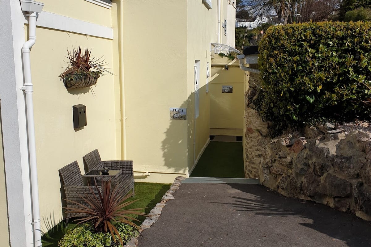 A narrow driveway leading to a building with a yellow exterior, a small outdoor seating area, and a stone wall on one side.