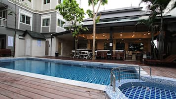 Uma piscina com deck de madeira, área de estar coberta ao ar livre e um edifício com várias janelas e fachada de tijolos.