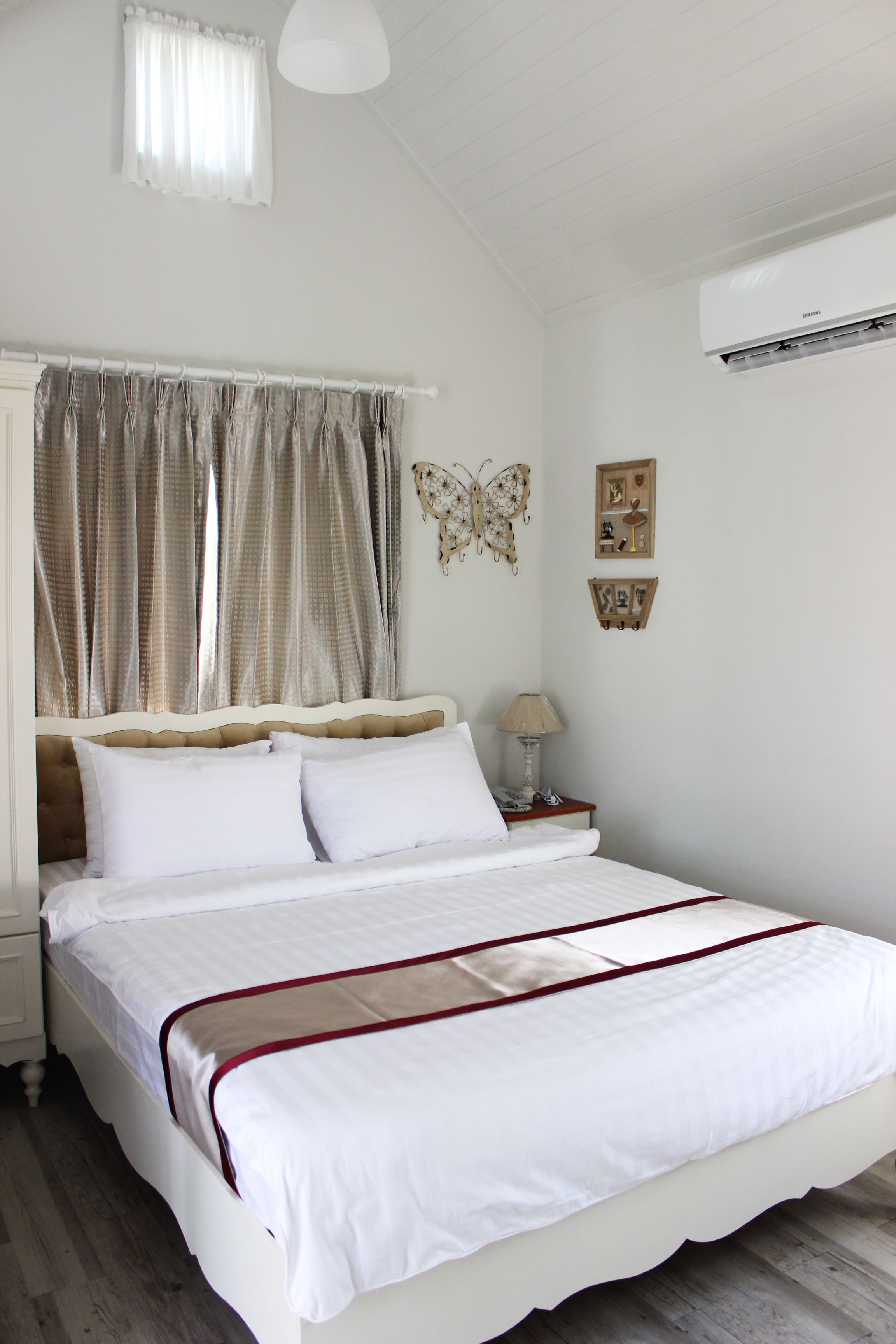 A neatly made bed with white bedding and a red trim, a bedside table with a lamp, and a wall-mounted air conditioner.