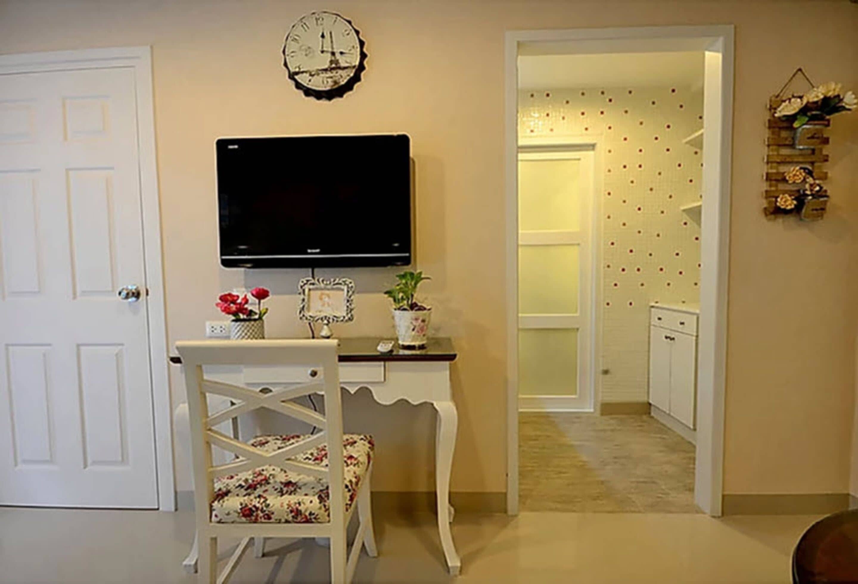 A room with a white desk, a television mounted above it, and a chair with floral upholstery. There is a door on the left and a hallway leading to a bathroom with a toilet and sink.