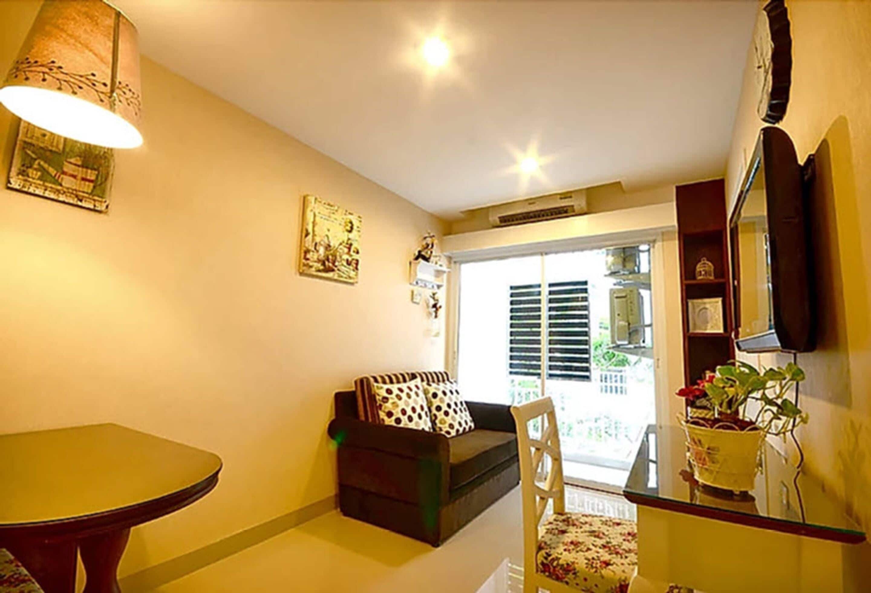 A cozy living room with a sofa, a dining table, and a chair. There is a window with blinds, a lamp, and a potted plant.