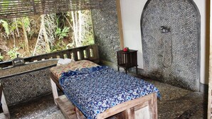 A room with a bed, a small table, a shower area with a pebble wall, and a window overlooking foliage. - Nefatari Exclusive Villas (Ubud)