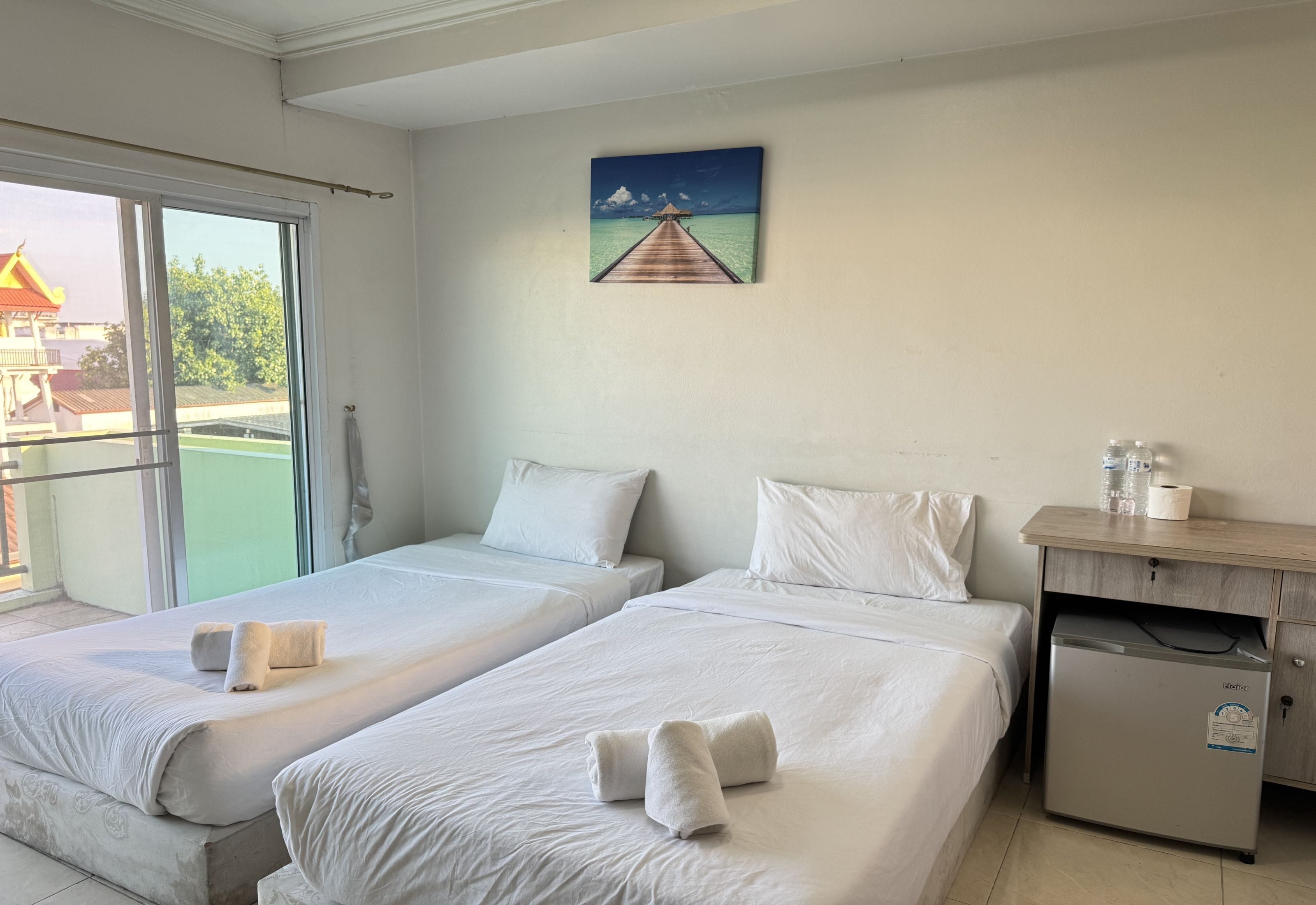 A hotel room with two beds, a small desk, a refrigerator, and a painting of a pier.