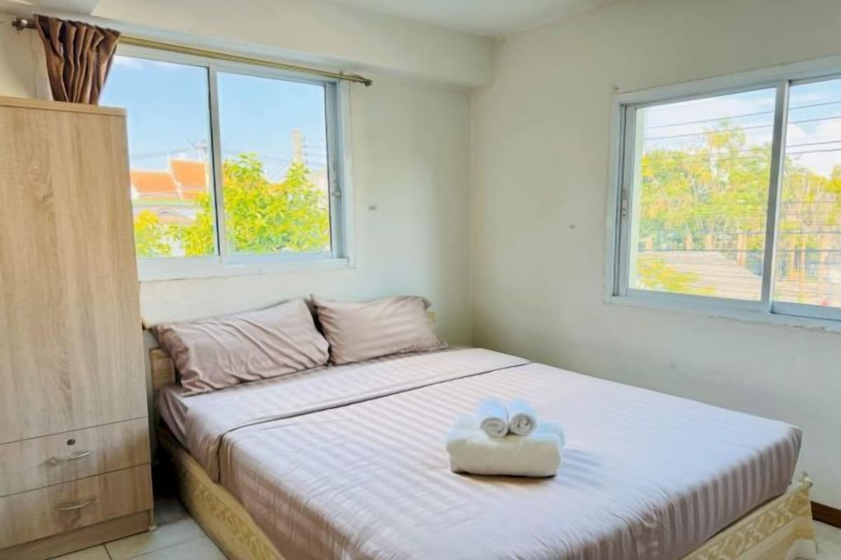 A bedroom with a bed, a wooden wardrobe, and two windows with a view of greenery.