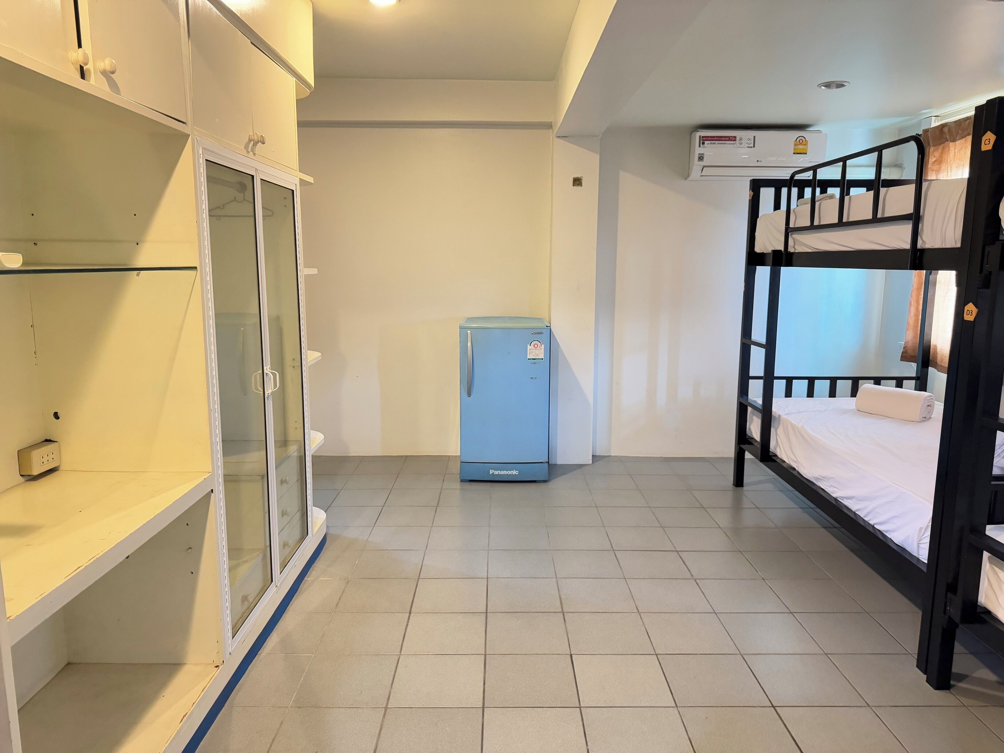 A dormitory room with a bunk bed, a small blue refrigerator, and a wardrobe with glass doors.