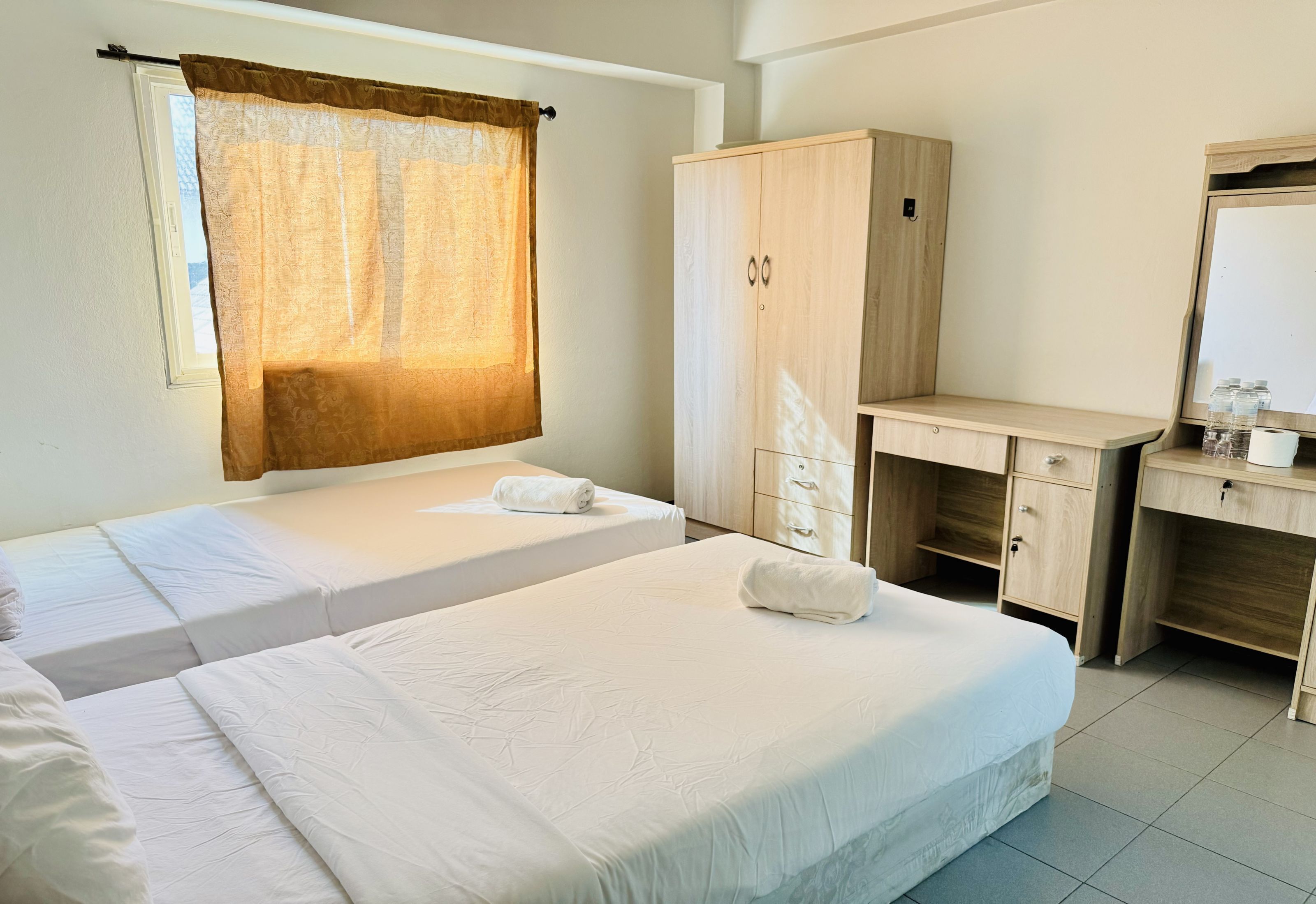 A hotel room with two single beds, a wooden wardrobe, a desk with a mirror, and a window with a sheer curtain.