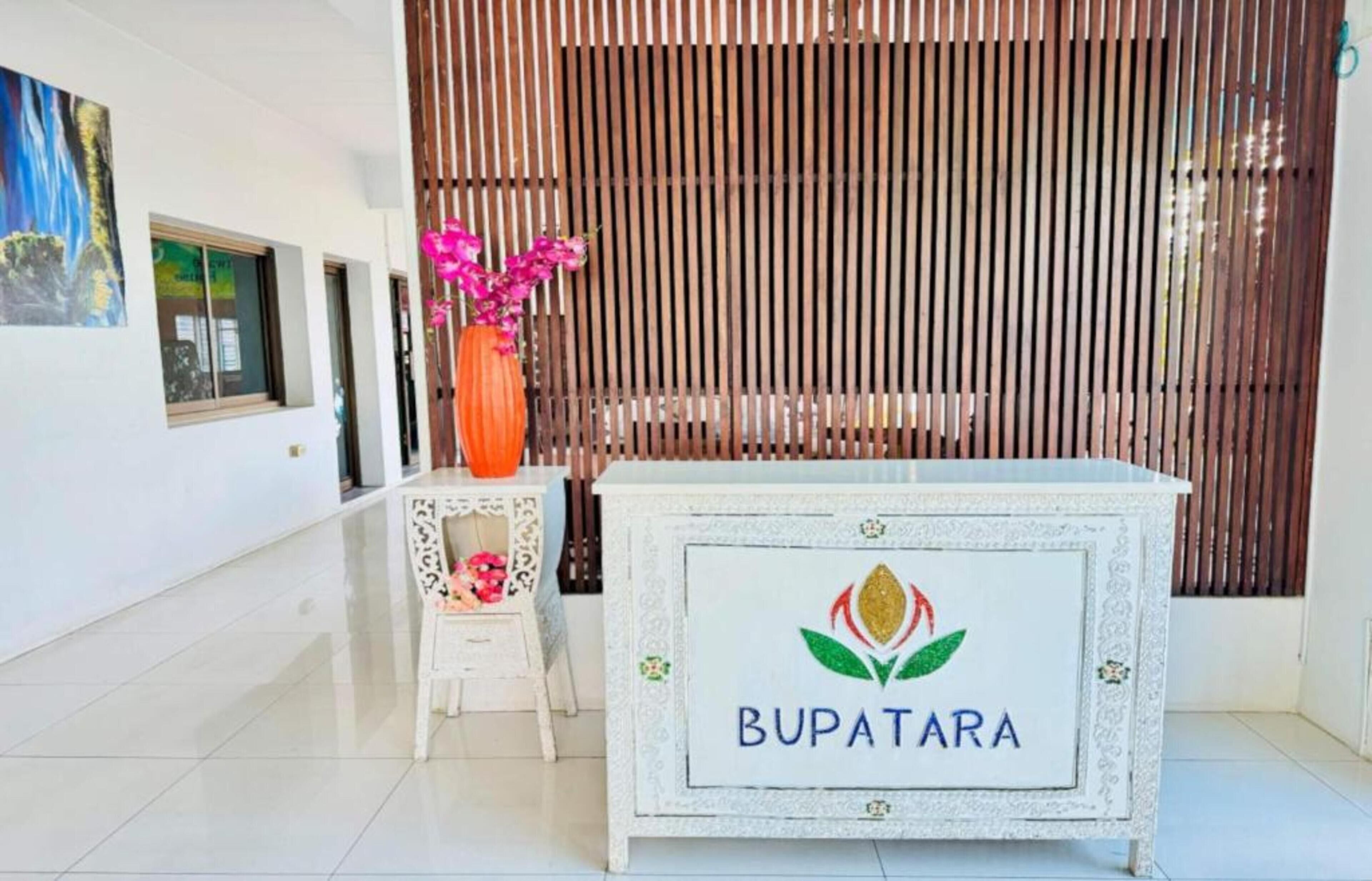 A reception area with a white reception desk labeled 'BUPATARA,' a small table with a vase of pink flowers, and a vertical wooden slat wall.