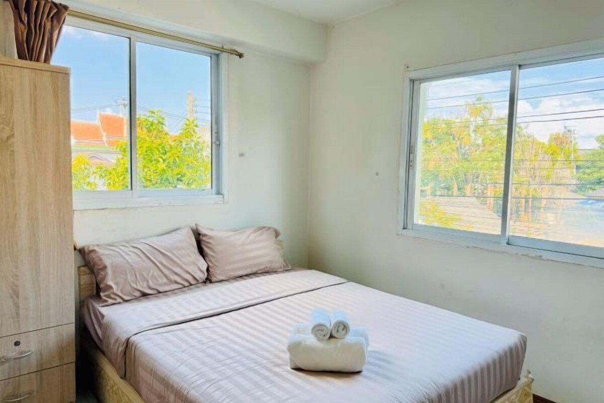 Una cama individual con una toalla doblada con esmero, un armario de madera y dos ventanas con vistas a zonas verdes y edificios.