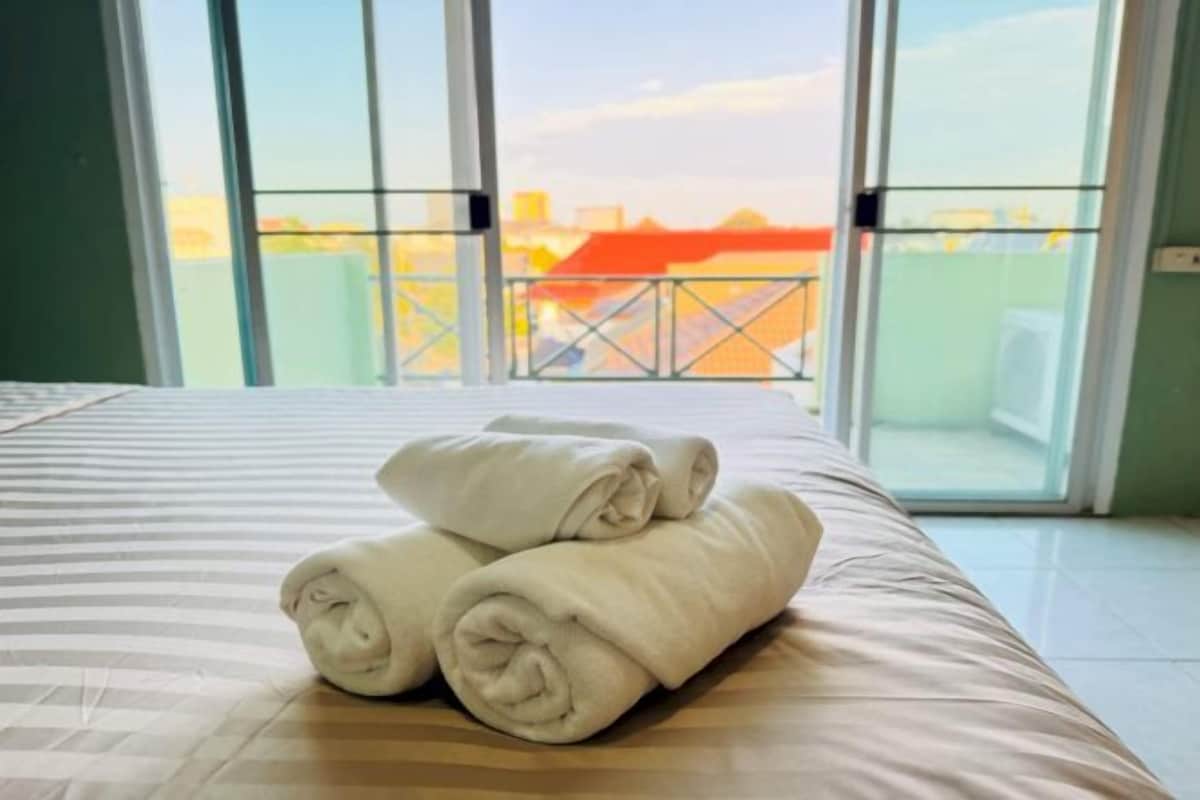 A hotel room with a bed, a view of a cityscape through large windows, and a balcony with a railing.