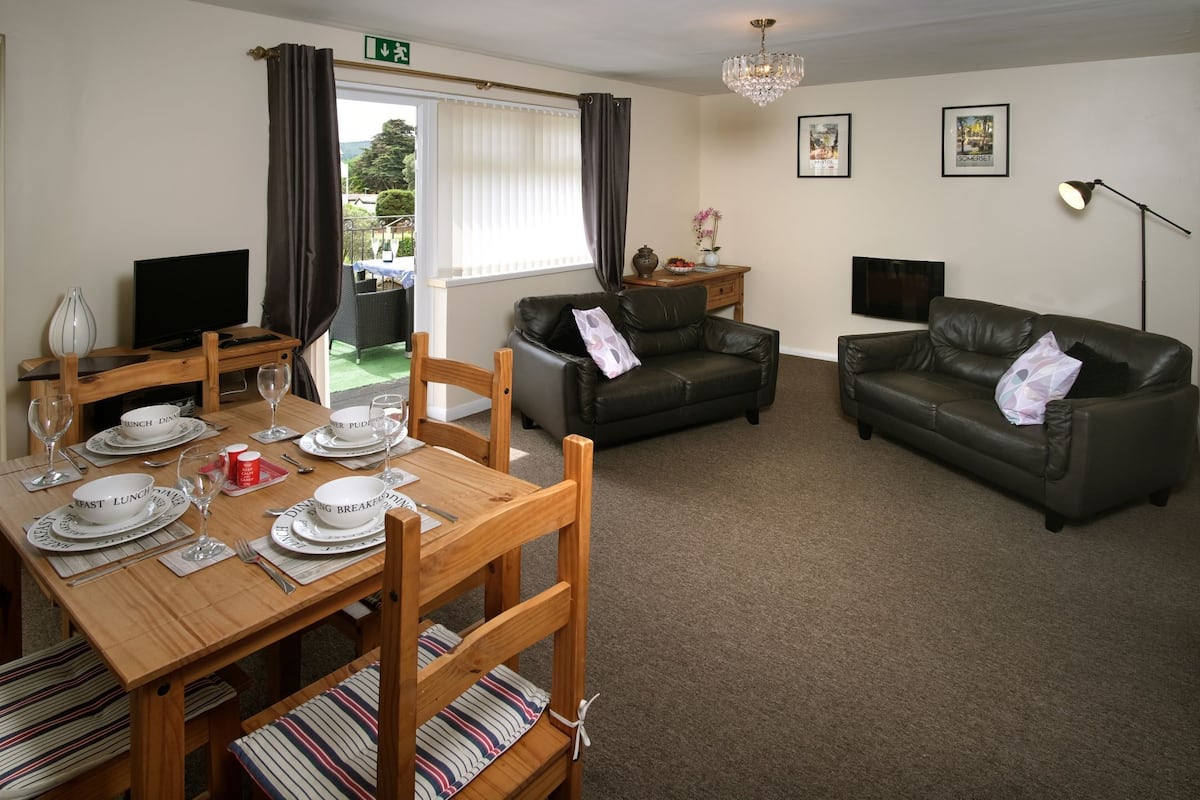 A furnished living room with a dining table set for breakfast, two leather sofas, a TV, and a chandelier.