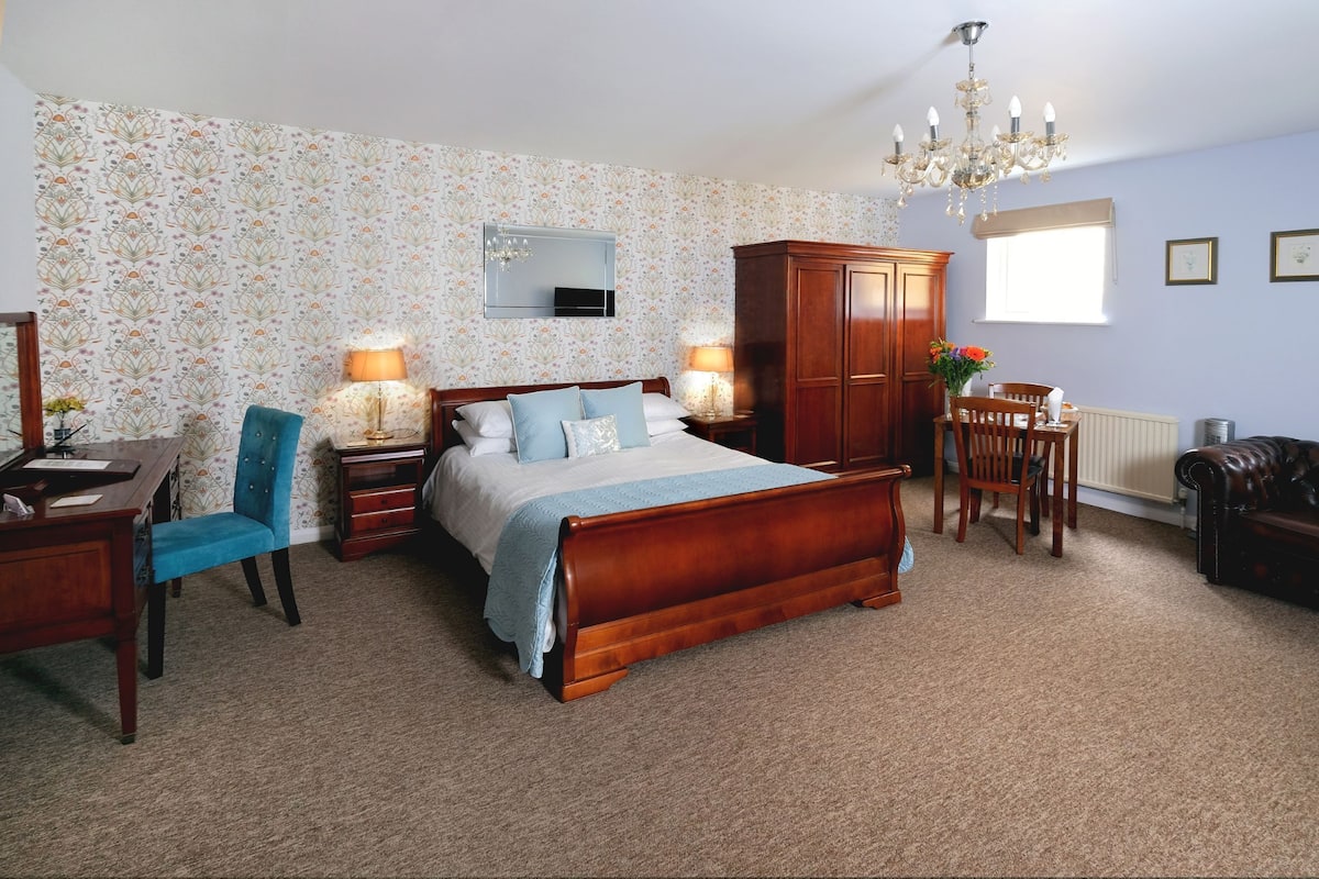A bedroom with a wooden bed, a blue chair, a desk, a dresser, a chandelier, and a window with blinds.